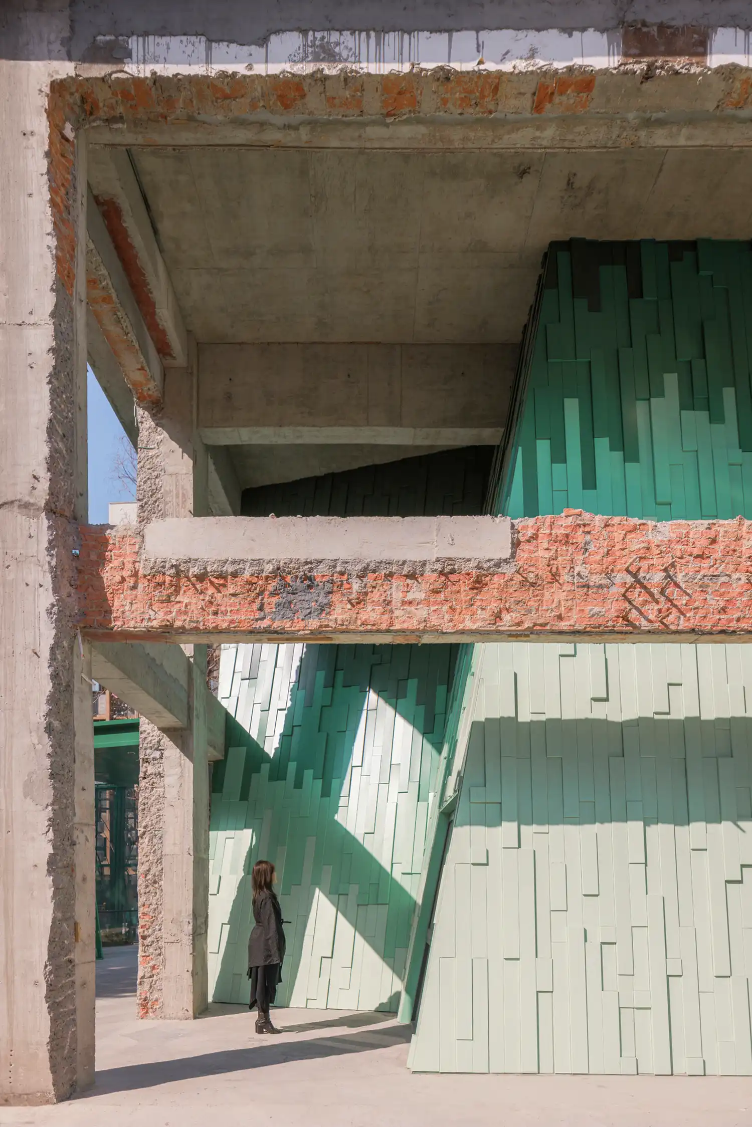 A person standing before the layered green metal mountain facade within the exposed concrete frame, highlighting the scale of the architectural installation.The "third way" of preservation: a visitor stands amidst the dialogue between Suzhou’s industrial past and the new, abstract landscape