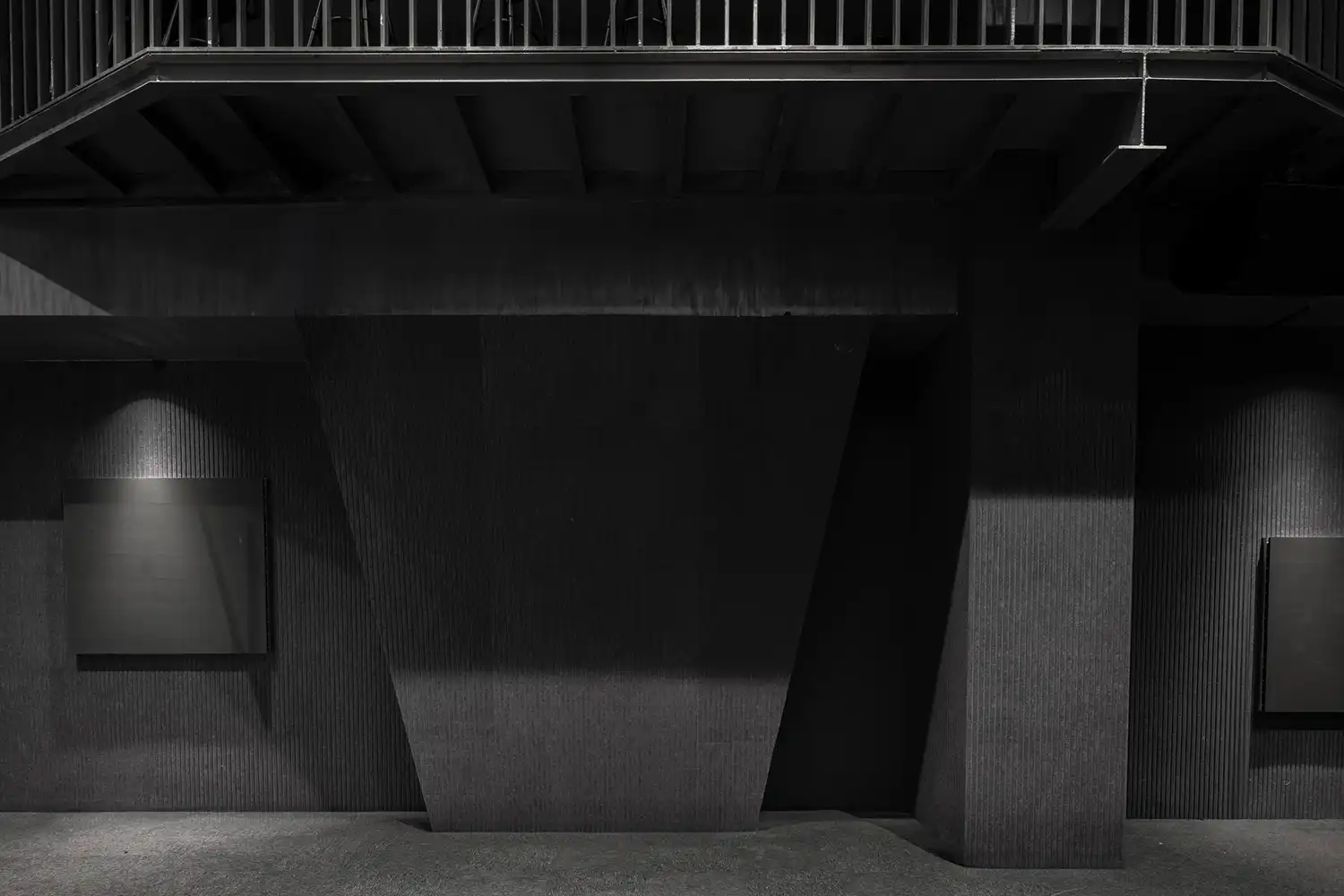 Minimalist black-on-black interior theater wall with vertical acoustic textures and a mezzanine level with a metal railing.