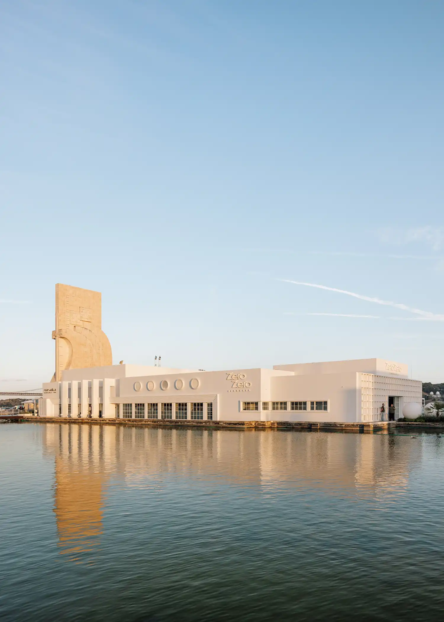 Exterior view of the white modernist Zero Zero Belém building reflected in a large pool at sunset.