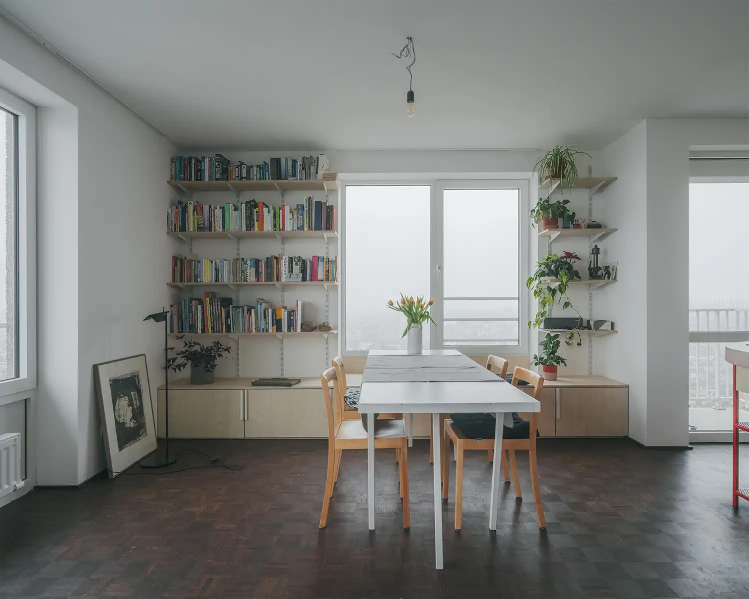 Minimalist dining area with white table, wooden chairs, and floor-to-ceiling bookshelves flanking a window.
