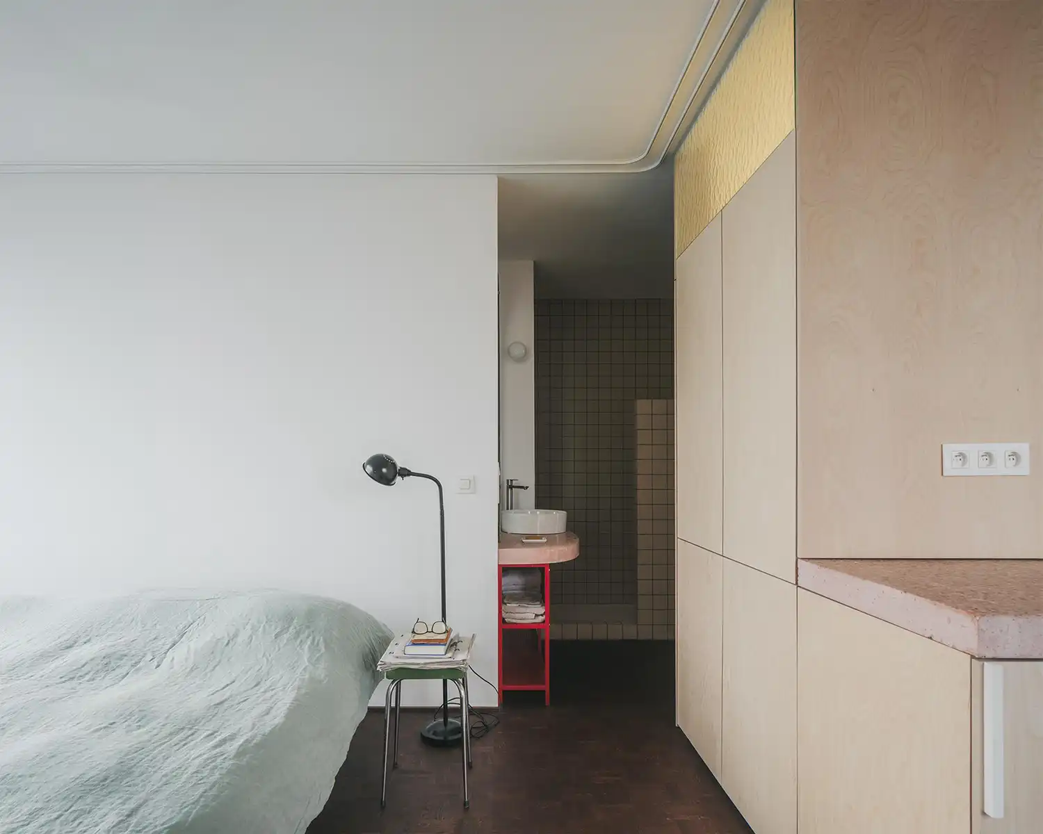 Minimalist bedroom area with green bedding next to a light wood wardrobe and pink stone vanity.