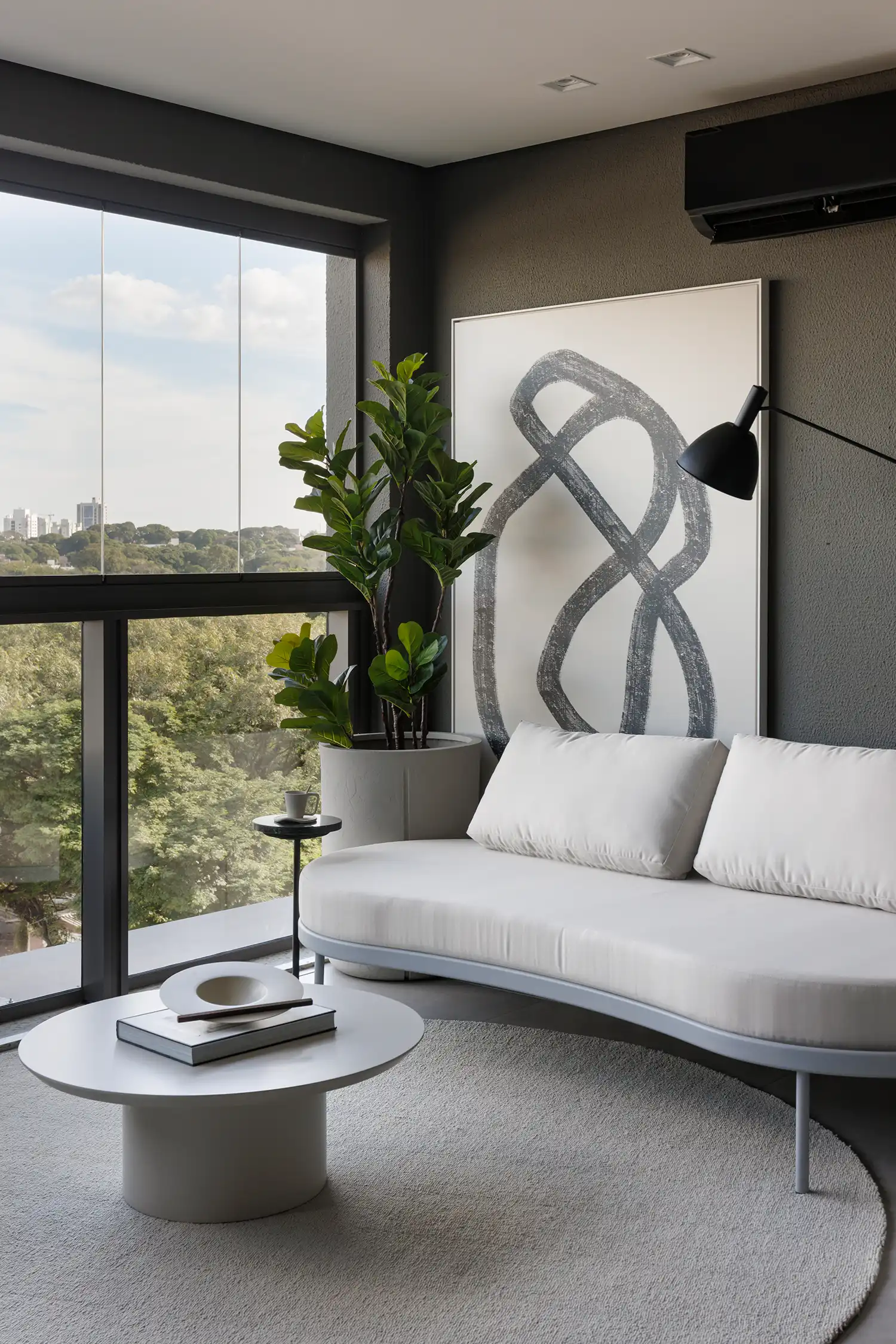 Modern balcony lounge area with a curved white sofa, a large plant, and a view of Maringá's greenery.
