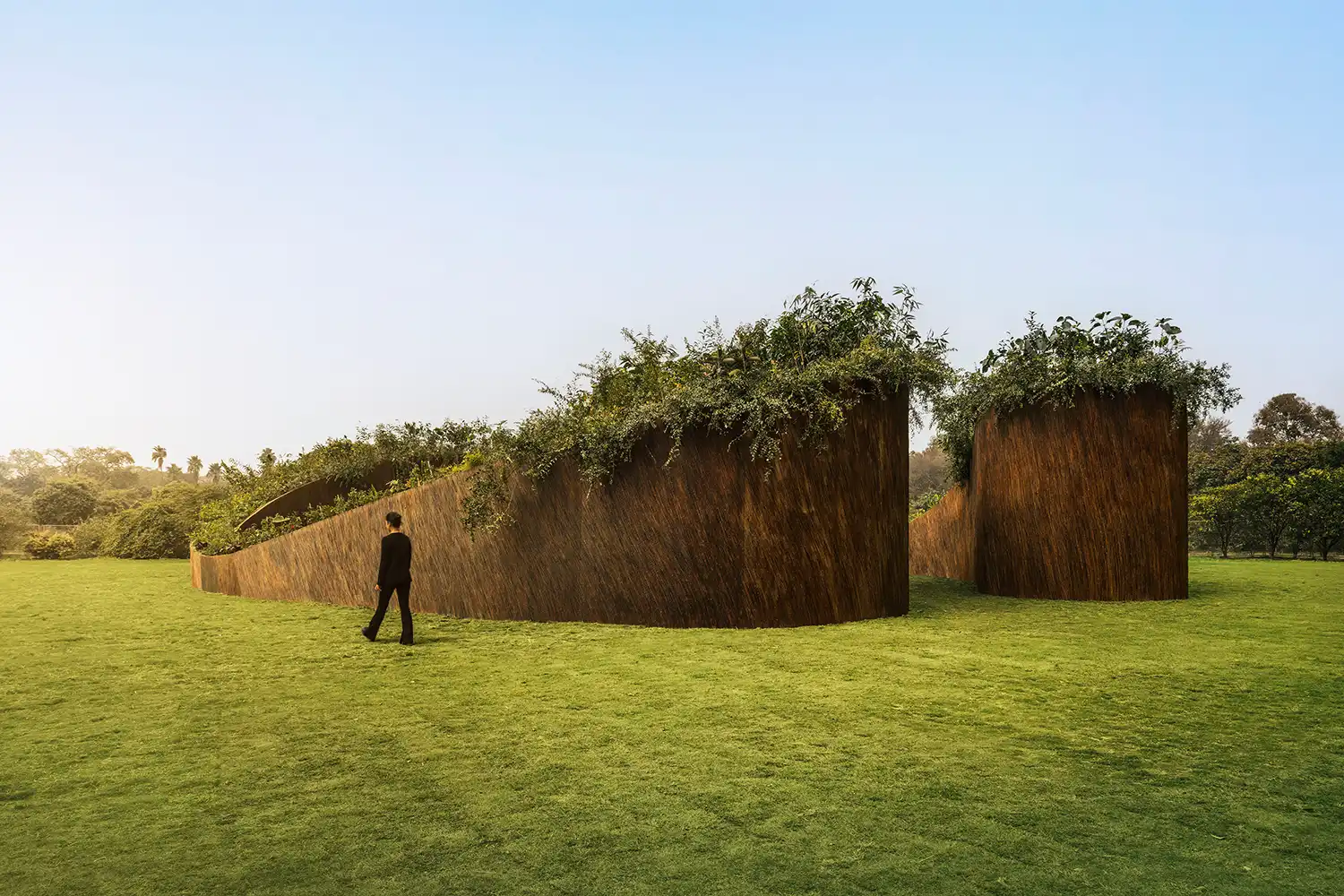 A person walking past the high, textured walls of the Aranyani Pavilion on a green lawn.