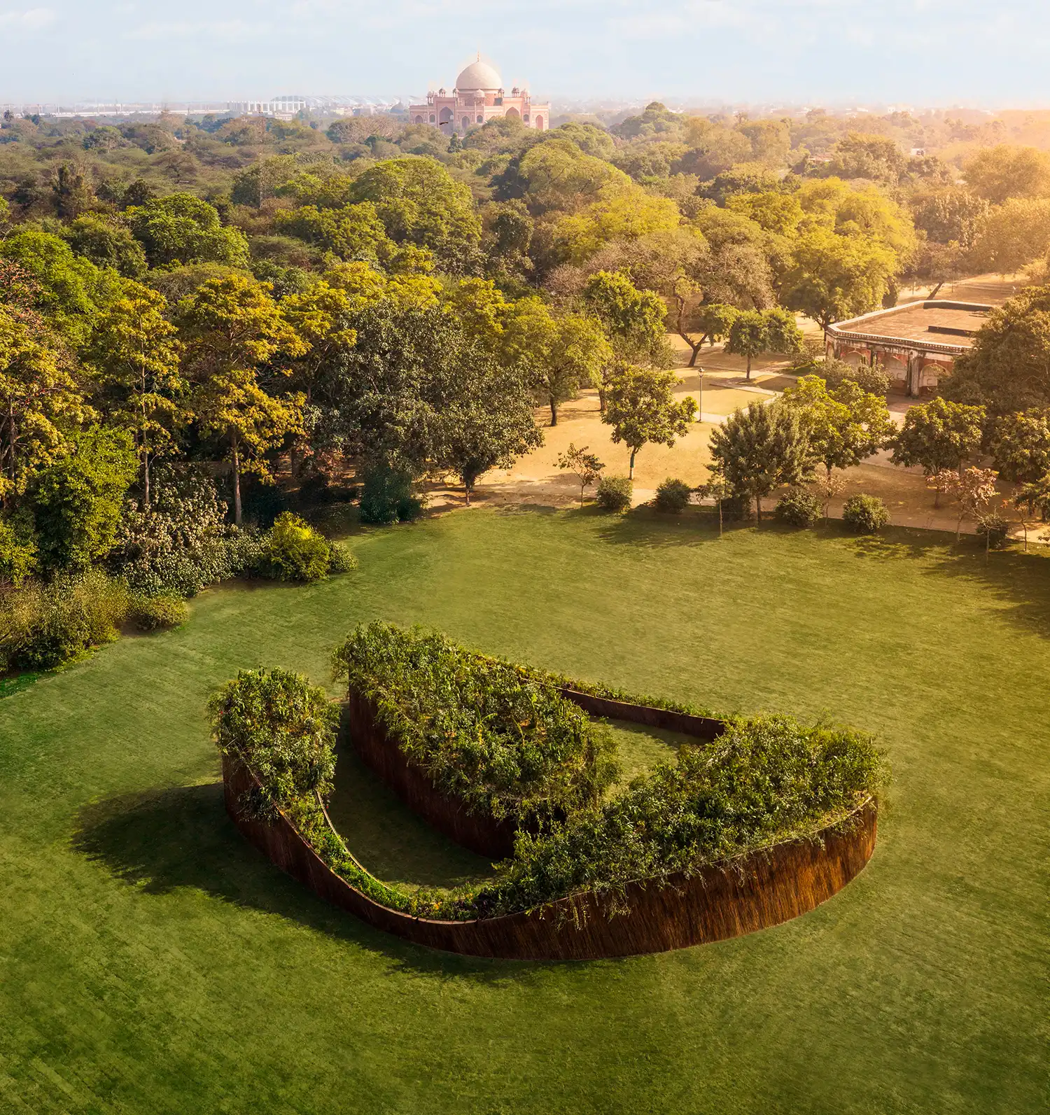 Bird's-eye view of the U-shaped Aranyani Pavilion in a vast park with a historic monument in the distance.