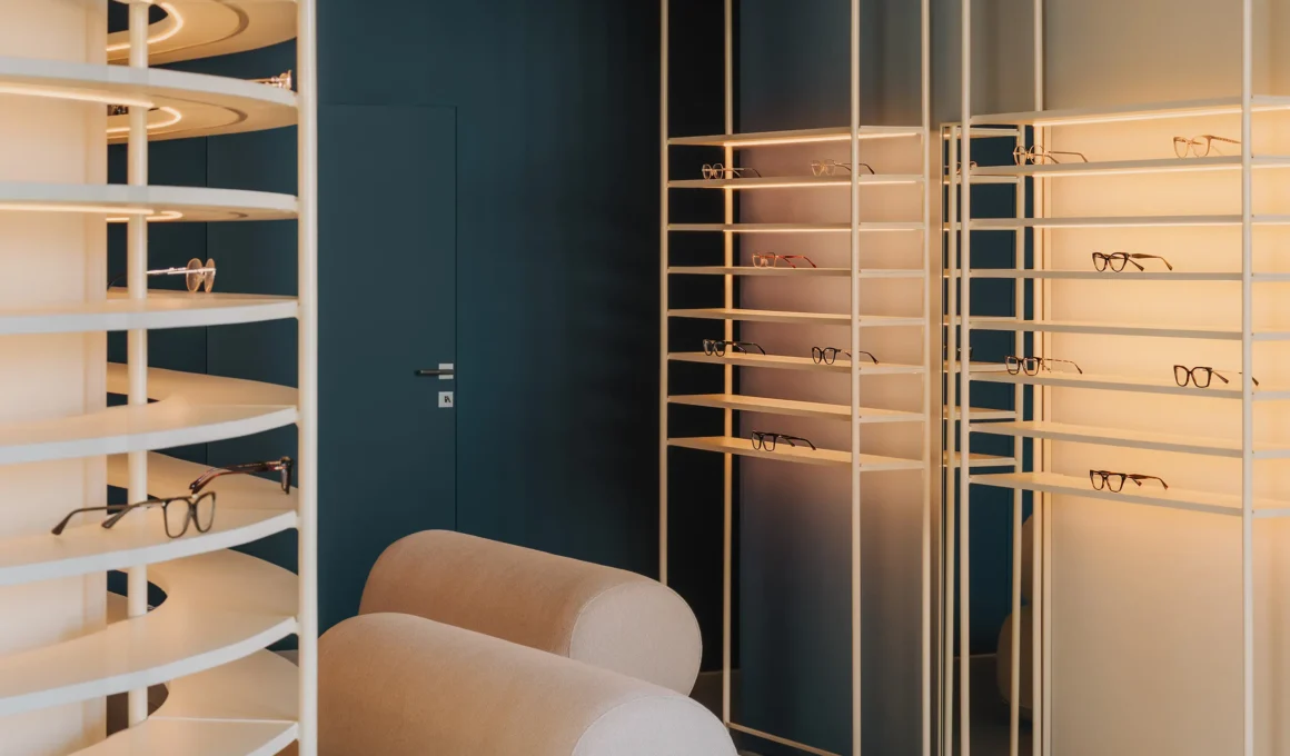 Interior detail of Auga Optical Store in Wrocław featuring curved white eyewear shelving, a textured glass partition, and deep navy walls.