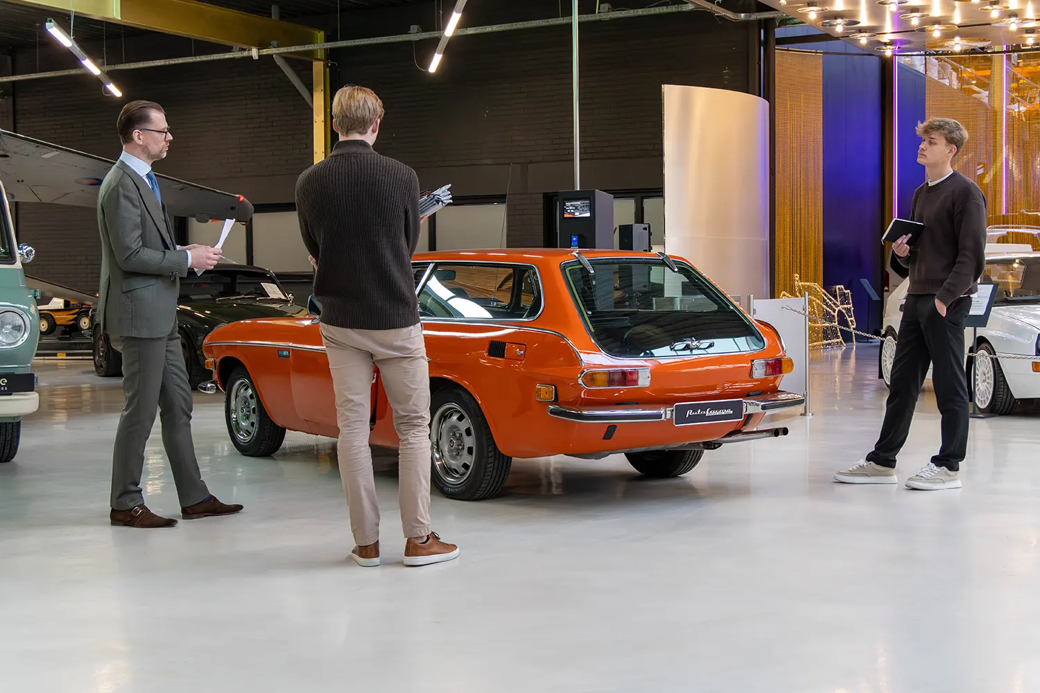 Design team and consultants reviewing an orange classic Volvo P1800 ES in a bright studio showroom.