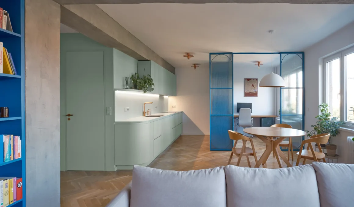 Open-plan living space in a Prague apartment with a sage green kitchen and blue industrial partitions.