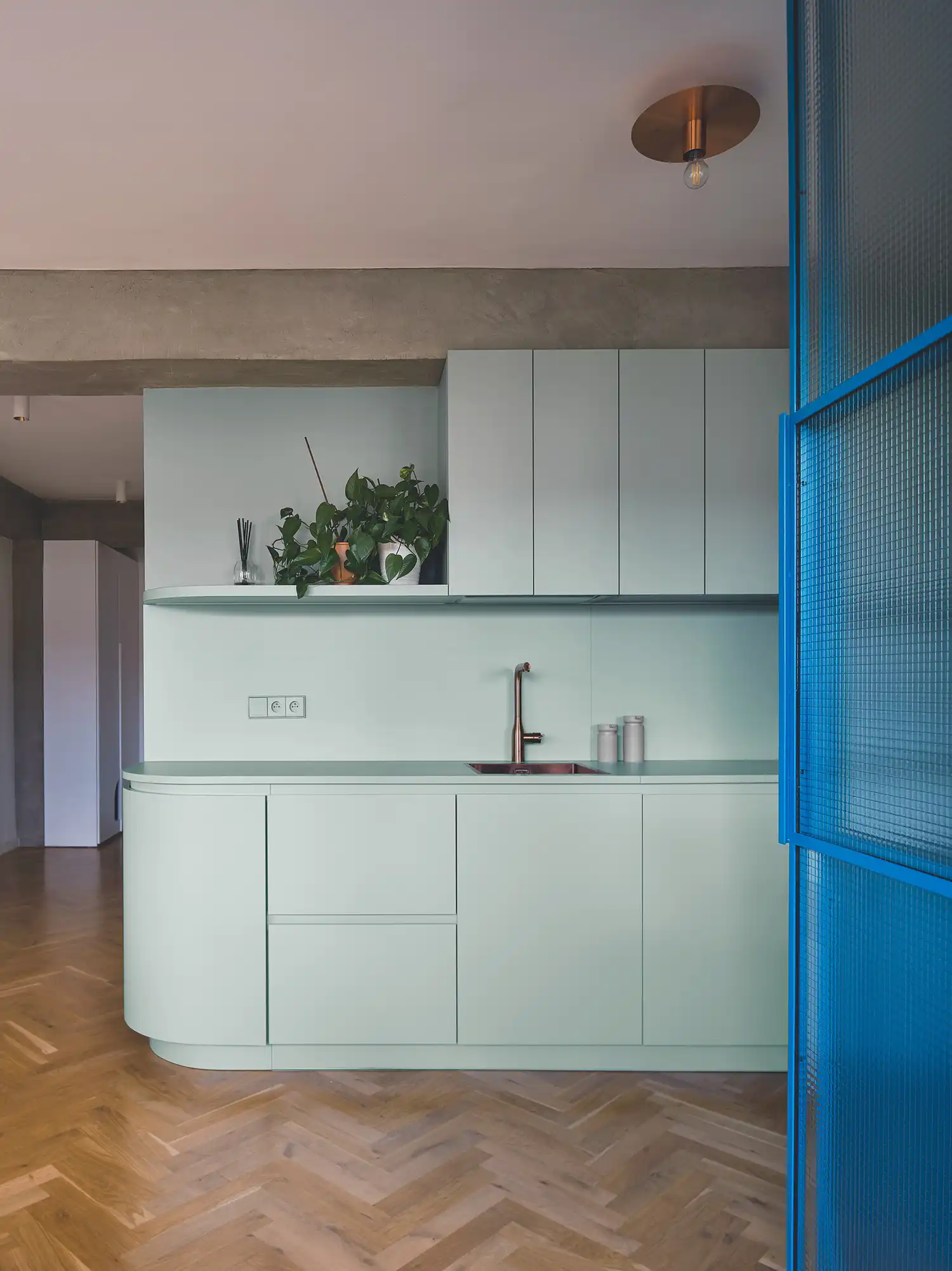 Close-up of a minimalist sage green kitchen with copper hardware and a concrete overhead beam.