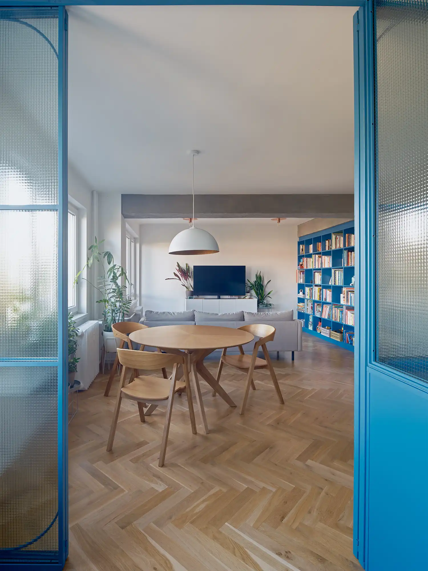 Dining area viewed through a blue fluted glass partition toward a grey sofa and TV unit.