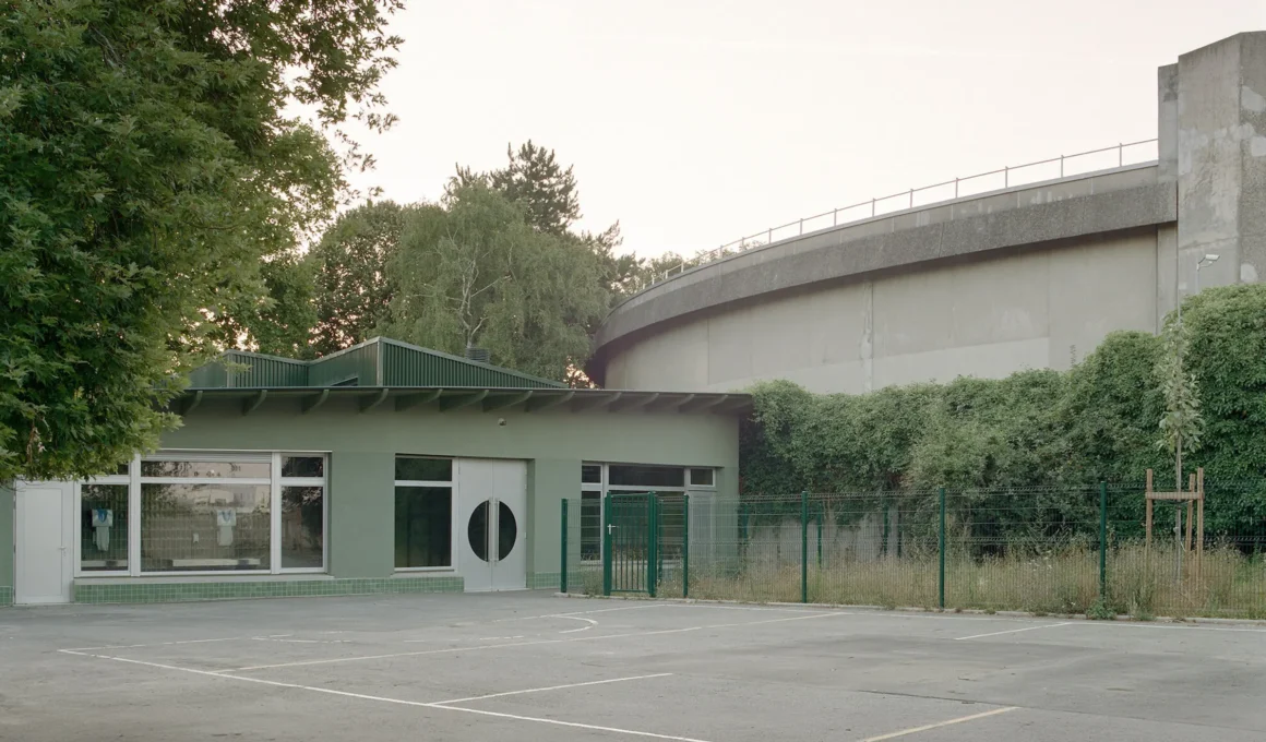 Exterior view of a single-story primary school building painted in sage green with green tiled baseboards, facing a playground.