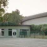 Exterior view of a single-story primary school building painted in sage green with green tiled baseboards, facing a playground.