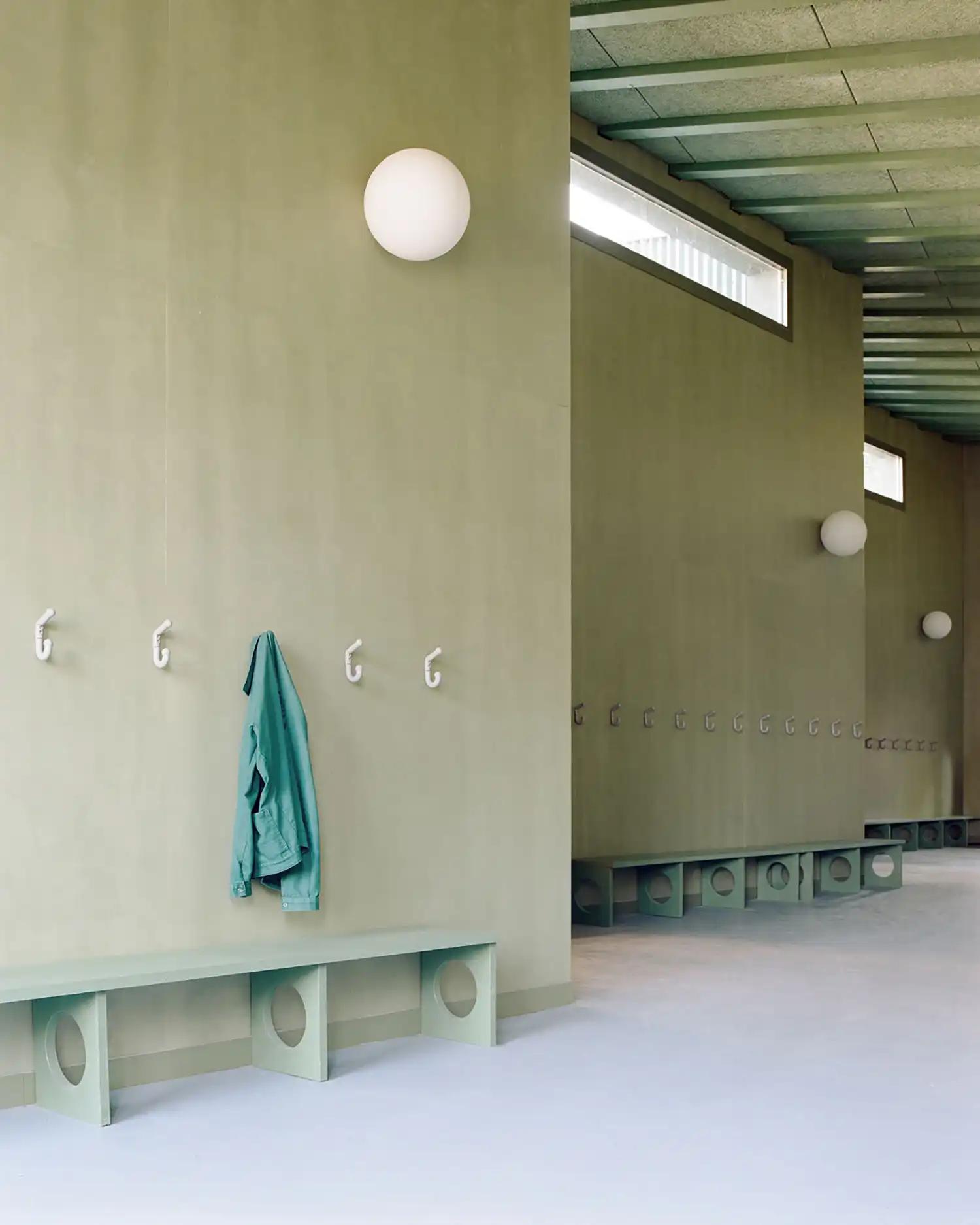 Interior view of a school corridor with muted green walls, white coat hooks, and simple benches with circular cutouts.