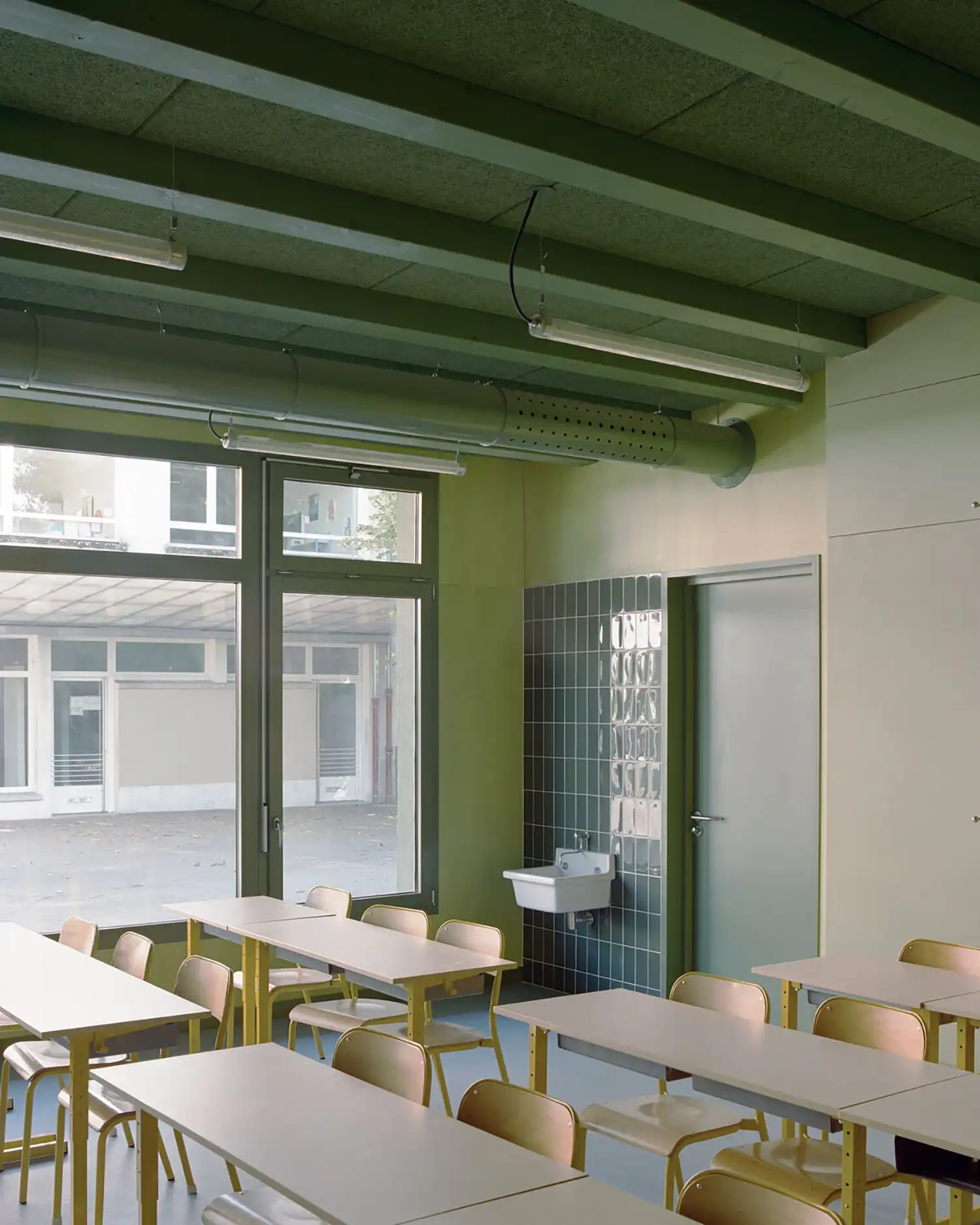 Inside a classroom with pale green walls, rows of student desks, and a small sink area tiled with dark green squares.