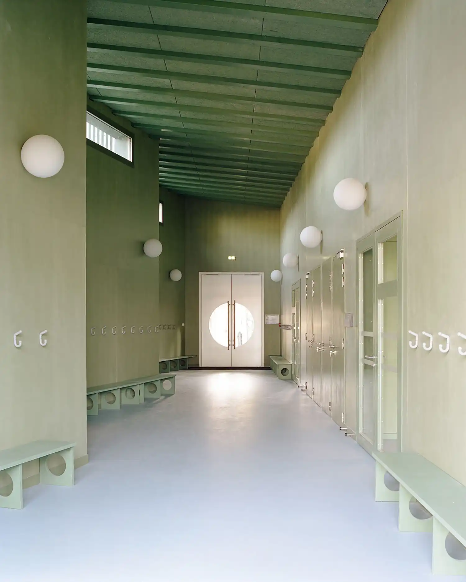 Long perspective shot of the school corridor looking towards a white double door with a circular window.