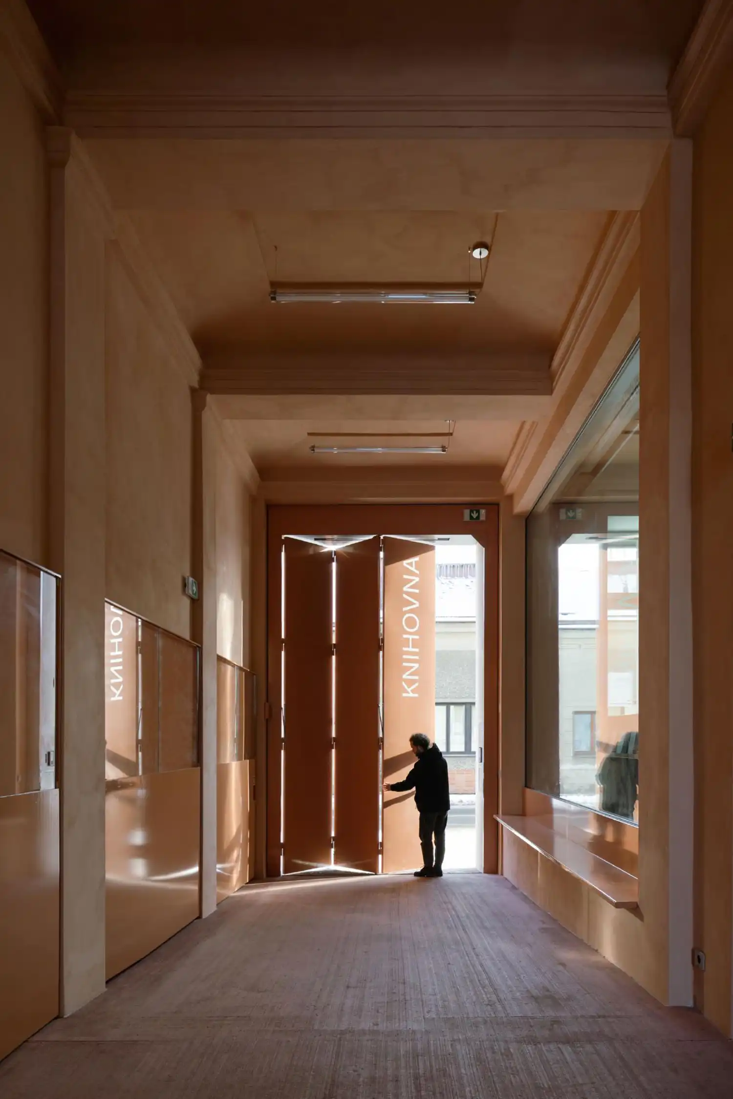 A person standing in a long, sunlit corridor with high ceilings and large pink folding doors at the end.