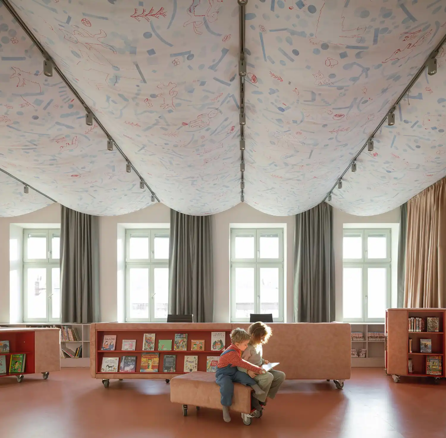 Two children sitting on a low pink bench in a library, reading a book together under a patterned canopy.