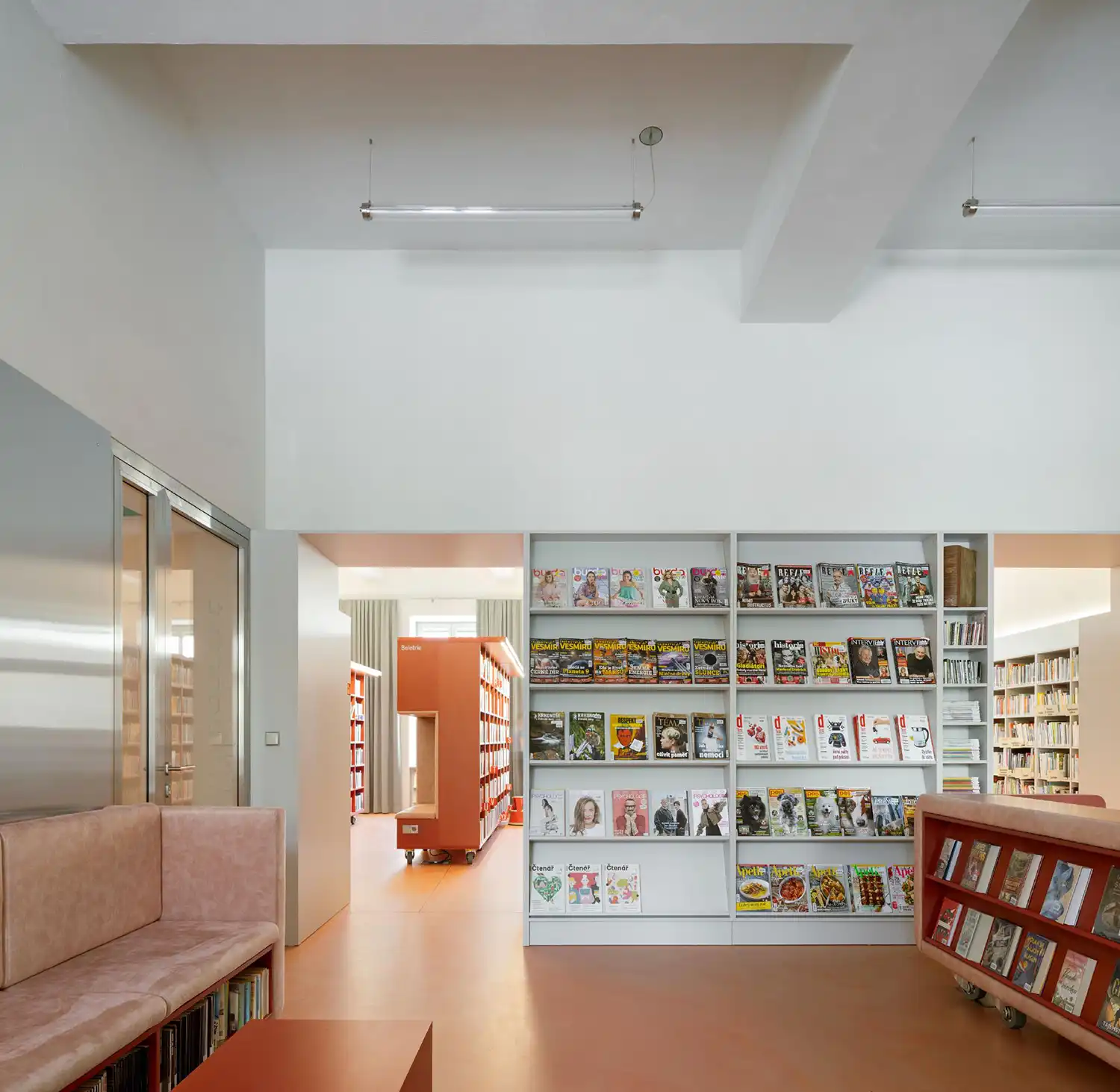 A clean, modern library interior with white walls, red flooring, and built-in shelving displaying magazines.