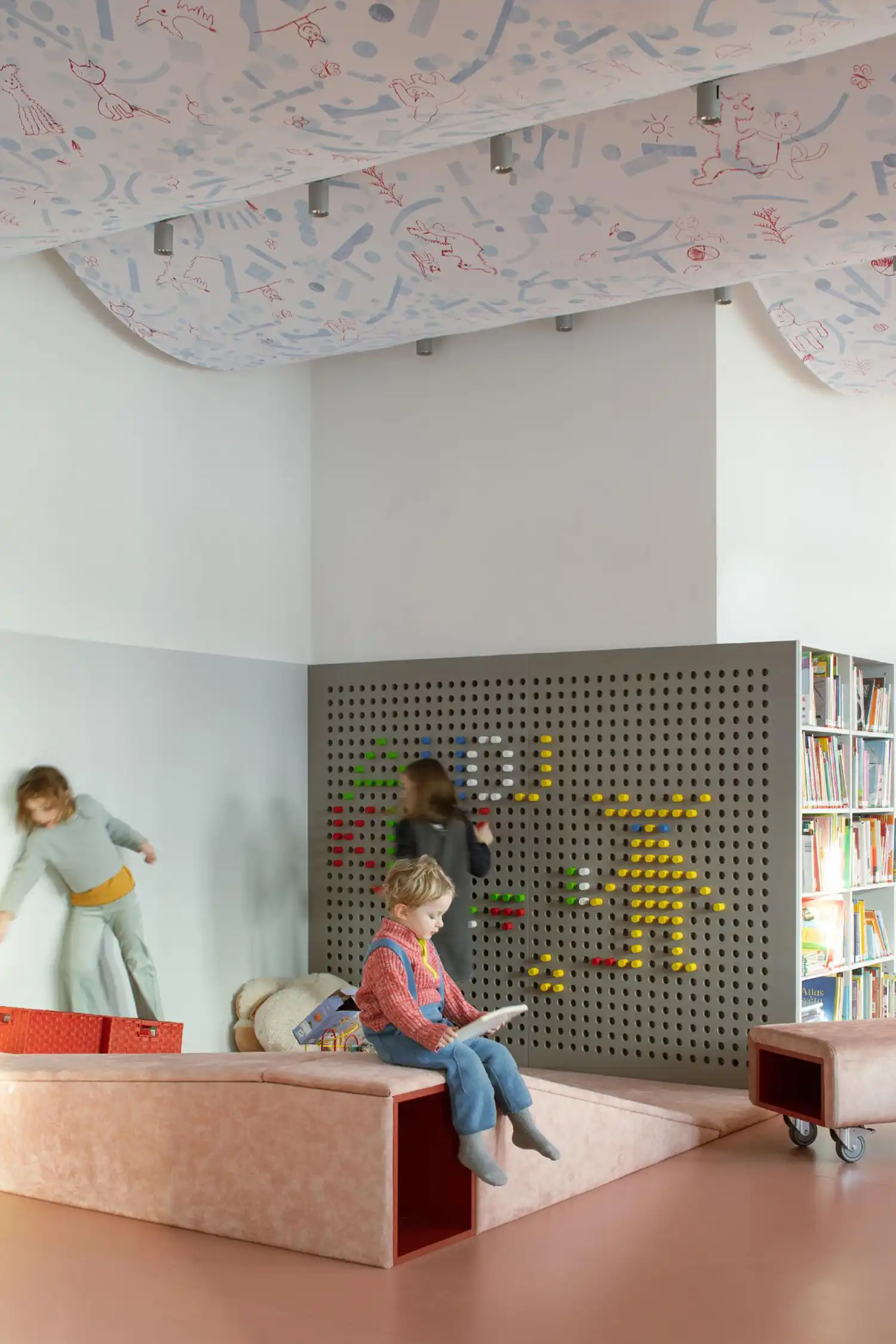 Children playing in a library corner featuring a large grey pegboard wall and soft pink play structures.
