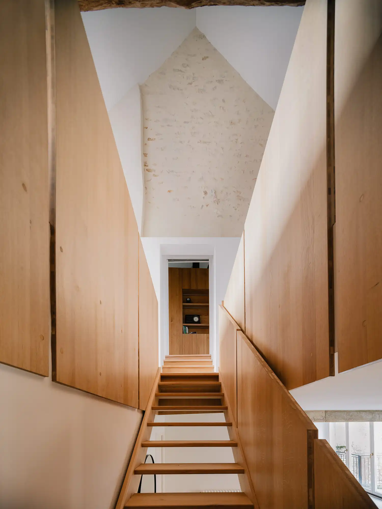 Looking up a narrow oak staircase toward a high vaulted ceiling with white plaster.