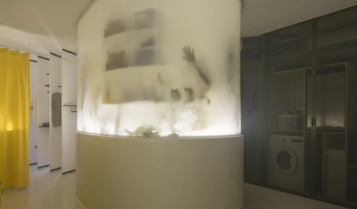 A glowing, translucent oval volume dominates the center of a minimalist room. The silhouette of a hand is visible through the polycarbonate skin, with shelves and plants blurred behind the material.