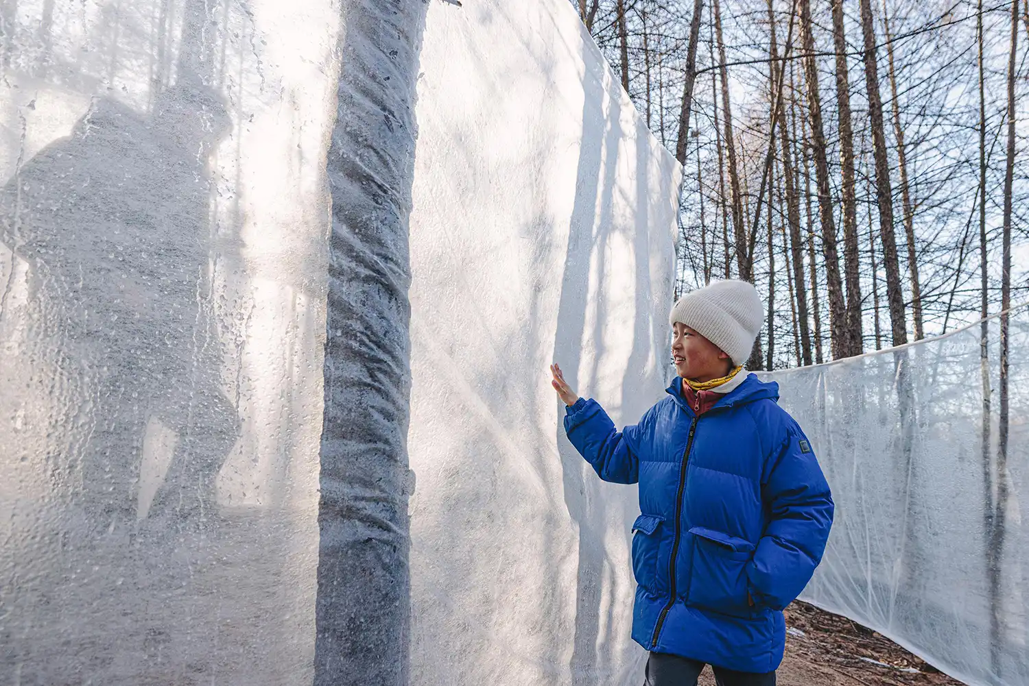 A young visitor in a blue jacket touching a thick, translucent ice wall with a silhouetted figure visible on the other side.