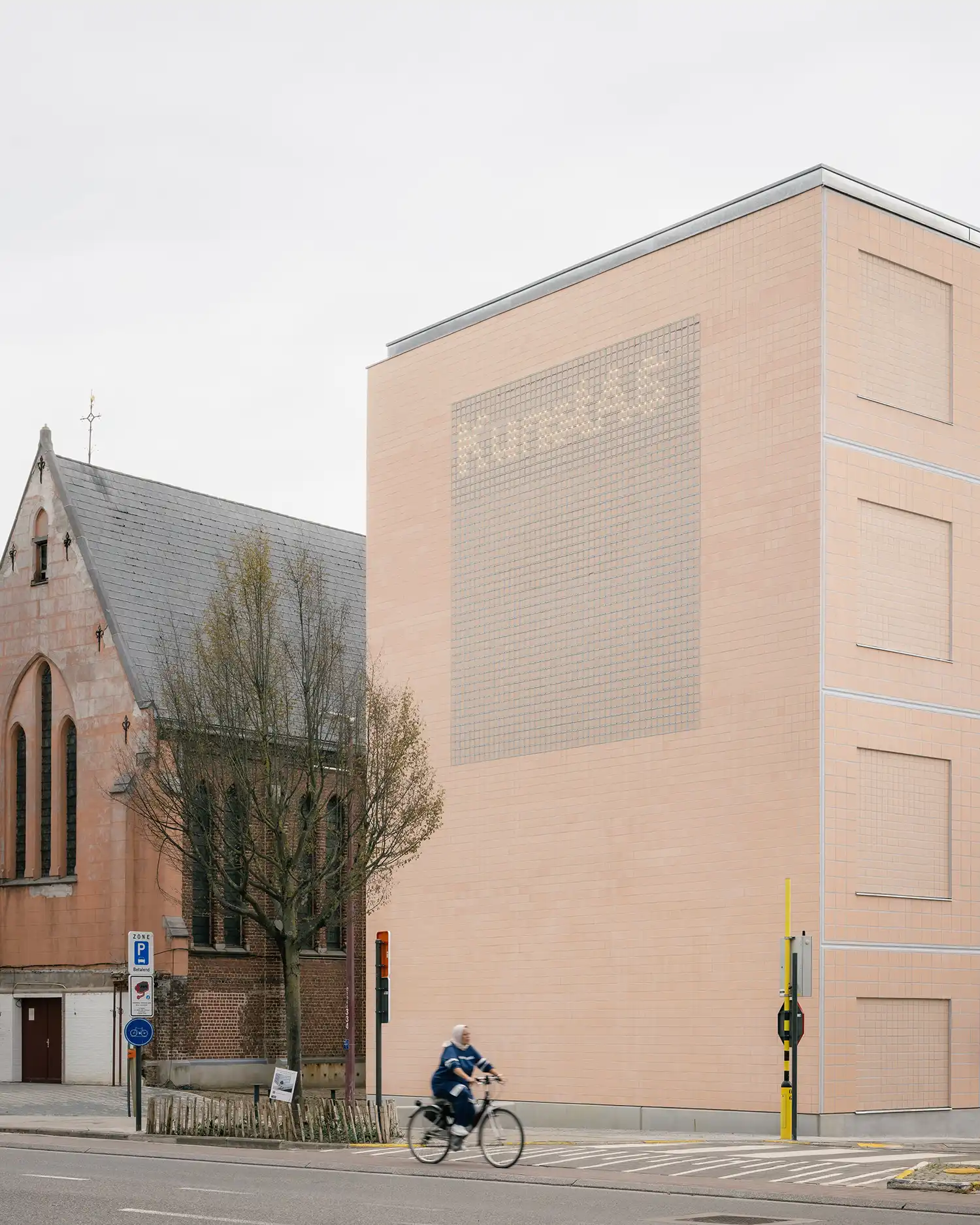 A tall, pastel-pink tiled facade of the Arts Campus next to a traditional brick chapel in Asse.