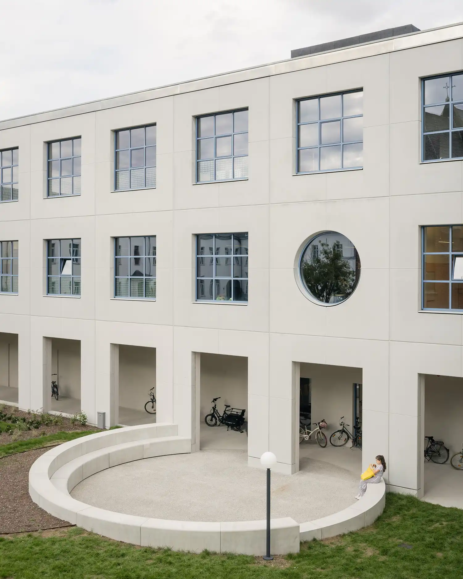A prefab concrete facade with a large circular window and a semi-circular concrete seating area in a garden.