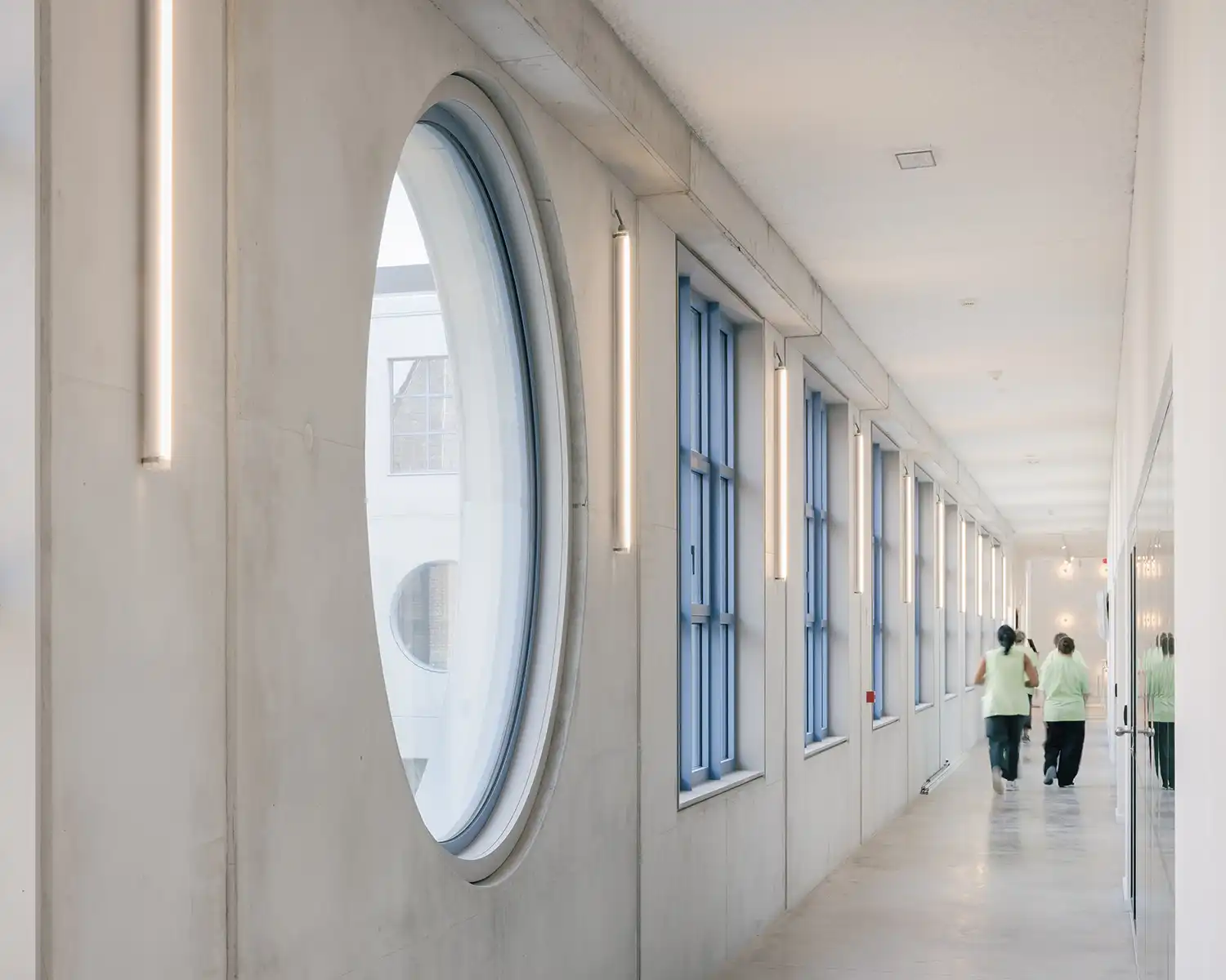 A long, bright interior corridor with large circular windows and blue-framed square windows.