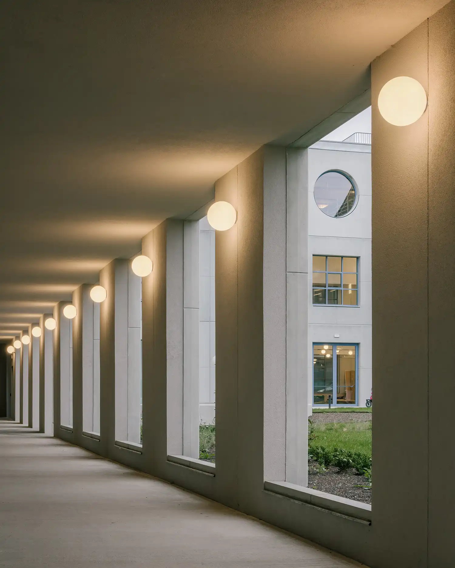 A covered concrete gallery with a row of glowing globe lights overlooking a green courtyard.