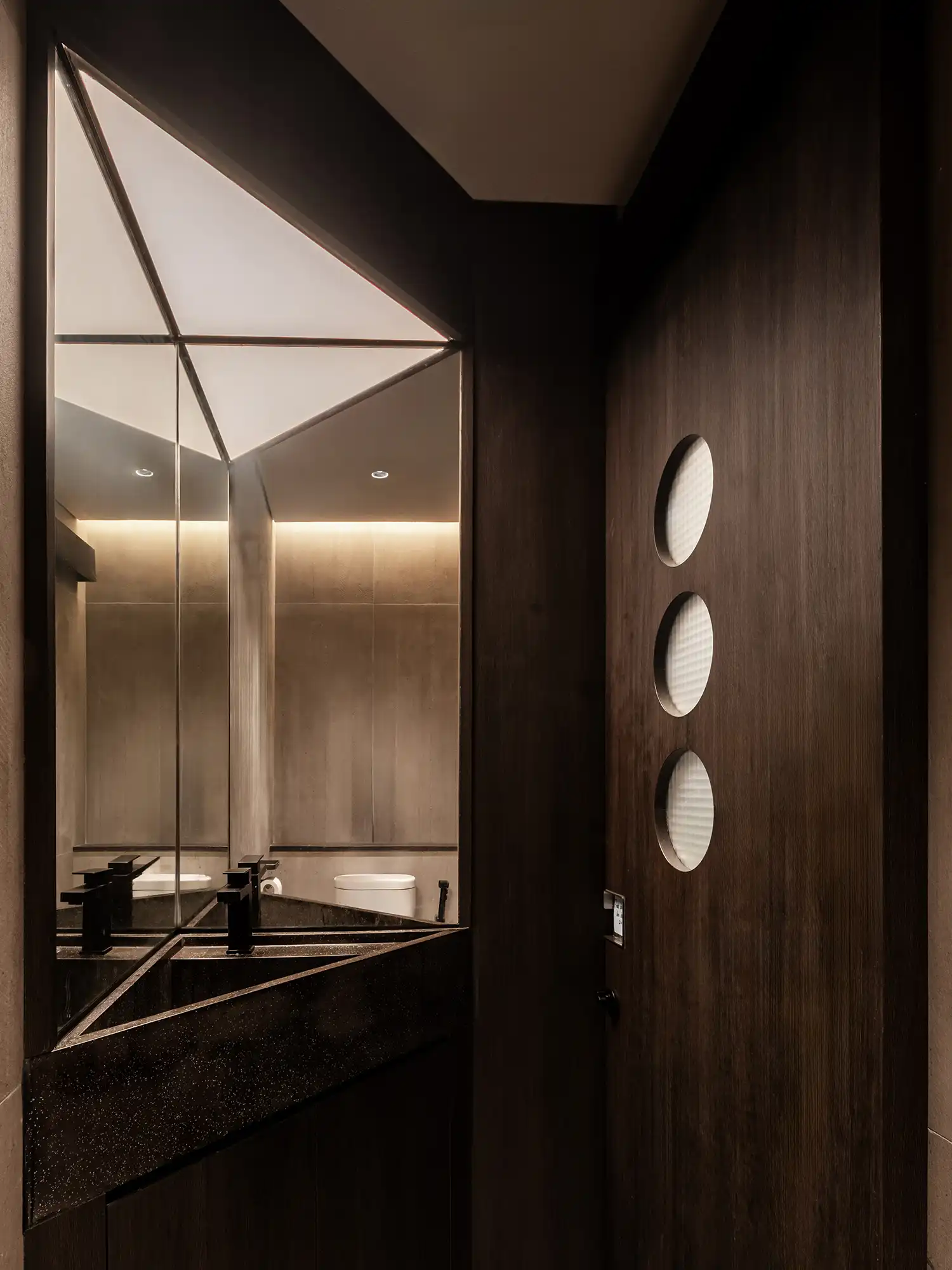 Minimalist dark wood restroom with a triangular illuminated mirror.