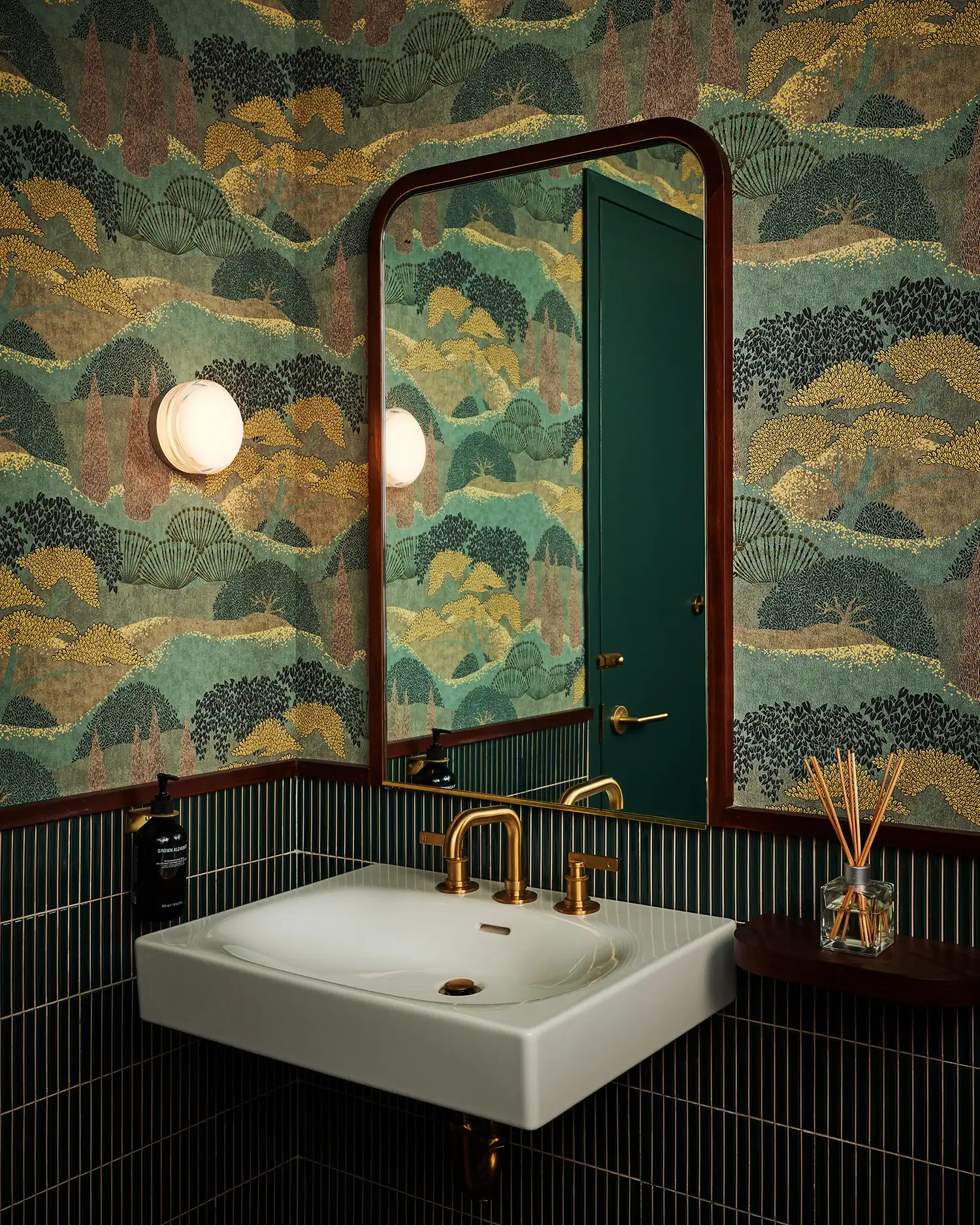 Modern bathroom with green vertical tiles, landscape wallpaper, and brass fixtures.