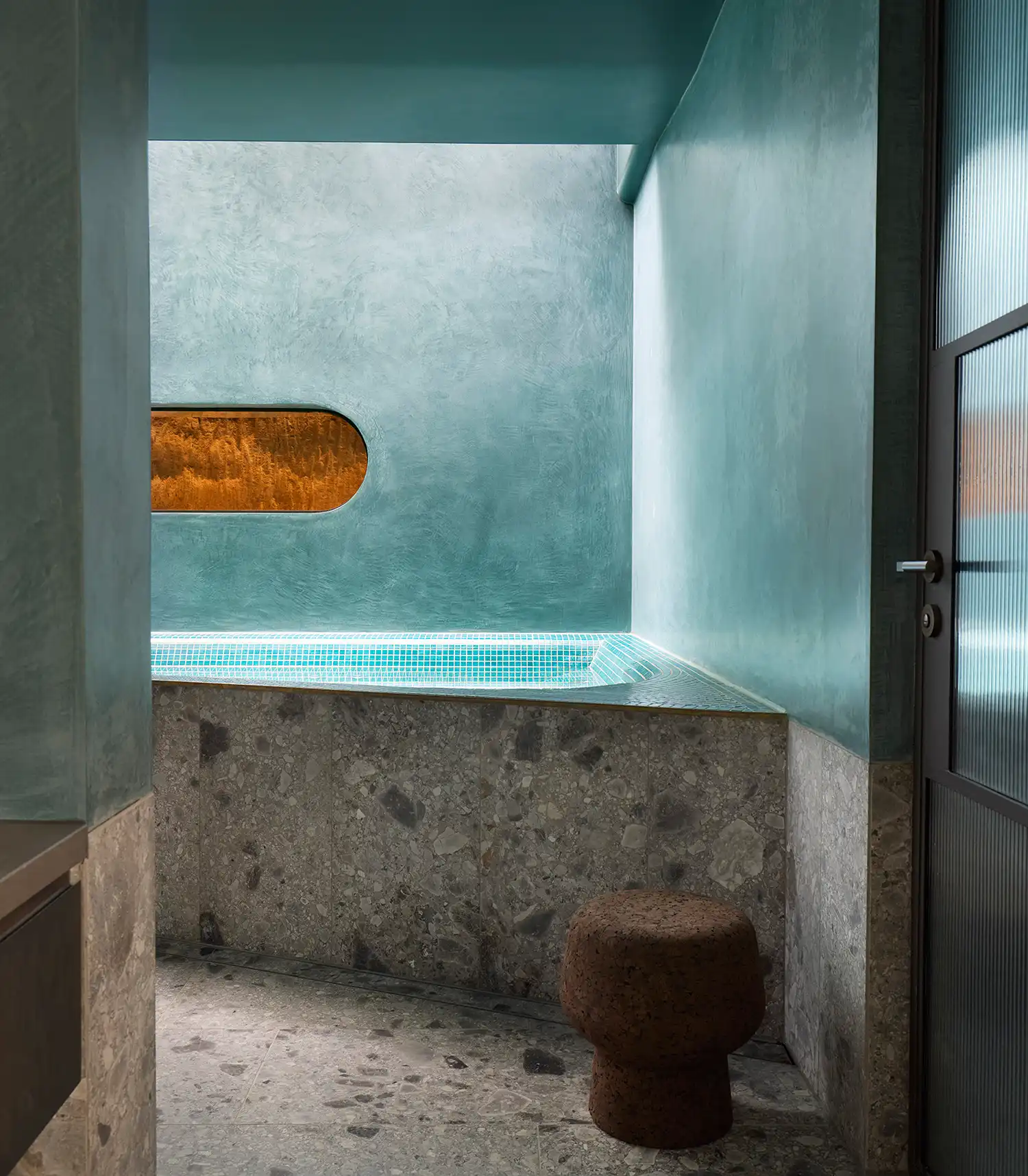 Minimalist spa room with a teal-colored wall, stone floor, and a small pool under natural light.