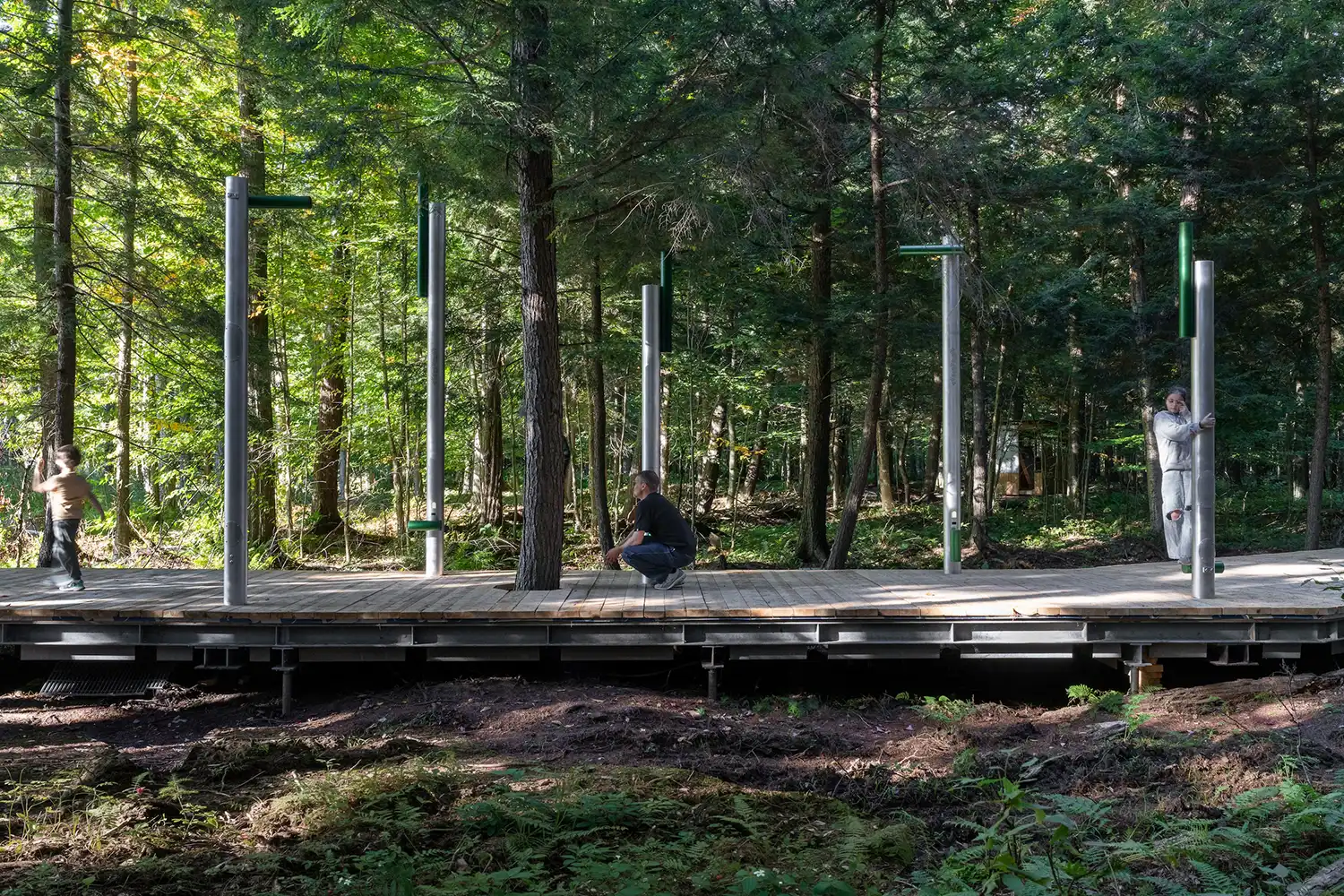 A side profile of the boardwalk showing humans interacting with the vertical aluminum poles amidst a dense canopy of trees.