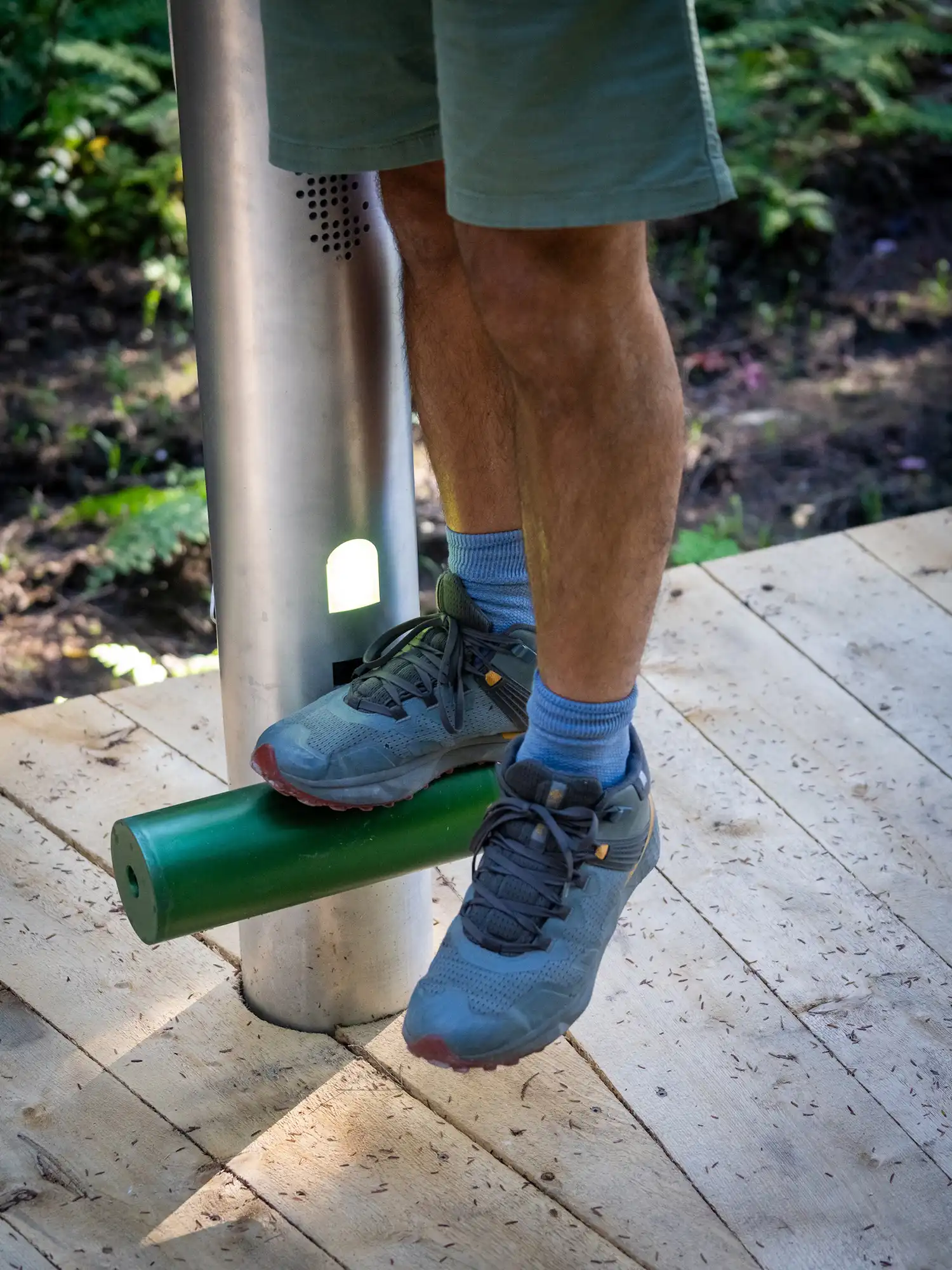 Macro view of a person’s feet in hiking shoes standing on a green perch, with a small light and speaker grille visible on the pole.