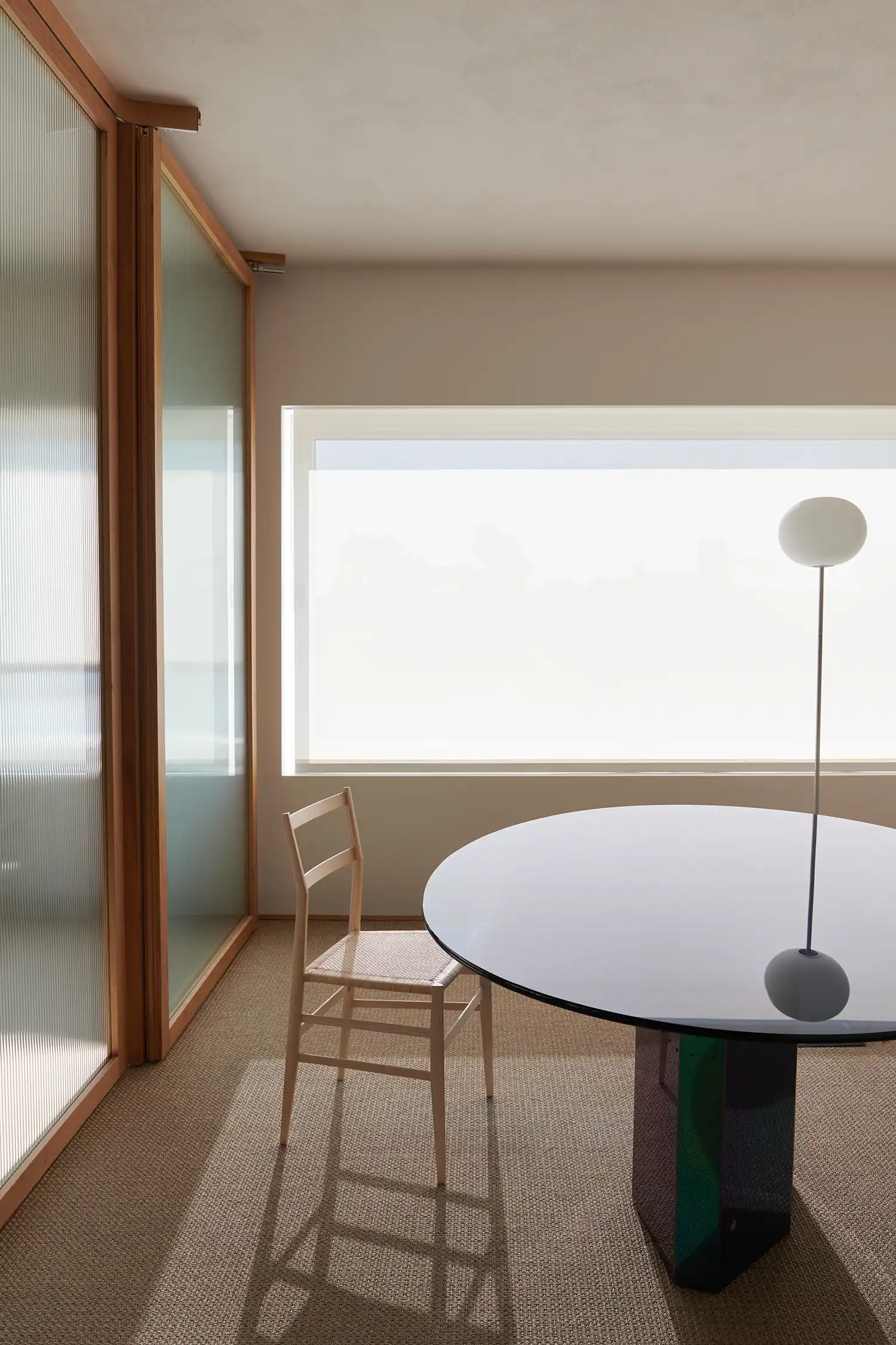 Minimalist dining area with a circular glass table, wooden chair, and ribbed glass partitions.