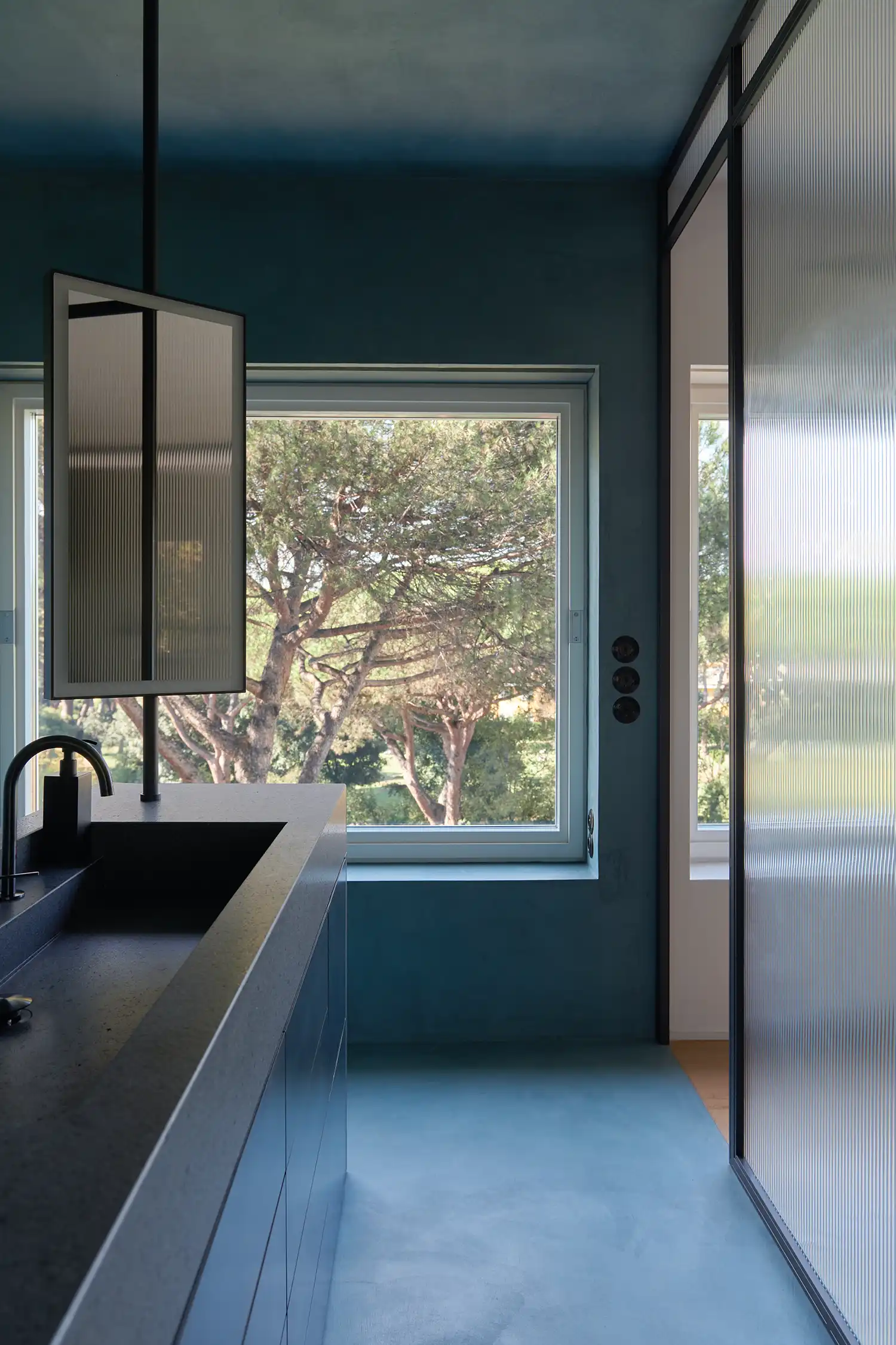 Modern blue bathroom with a stone basin, integrated mirror, and views of Roman pine trees.