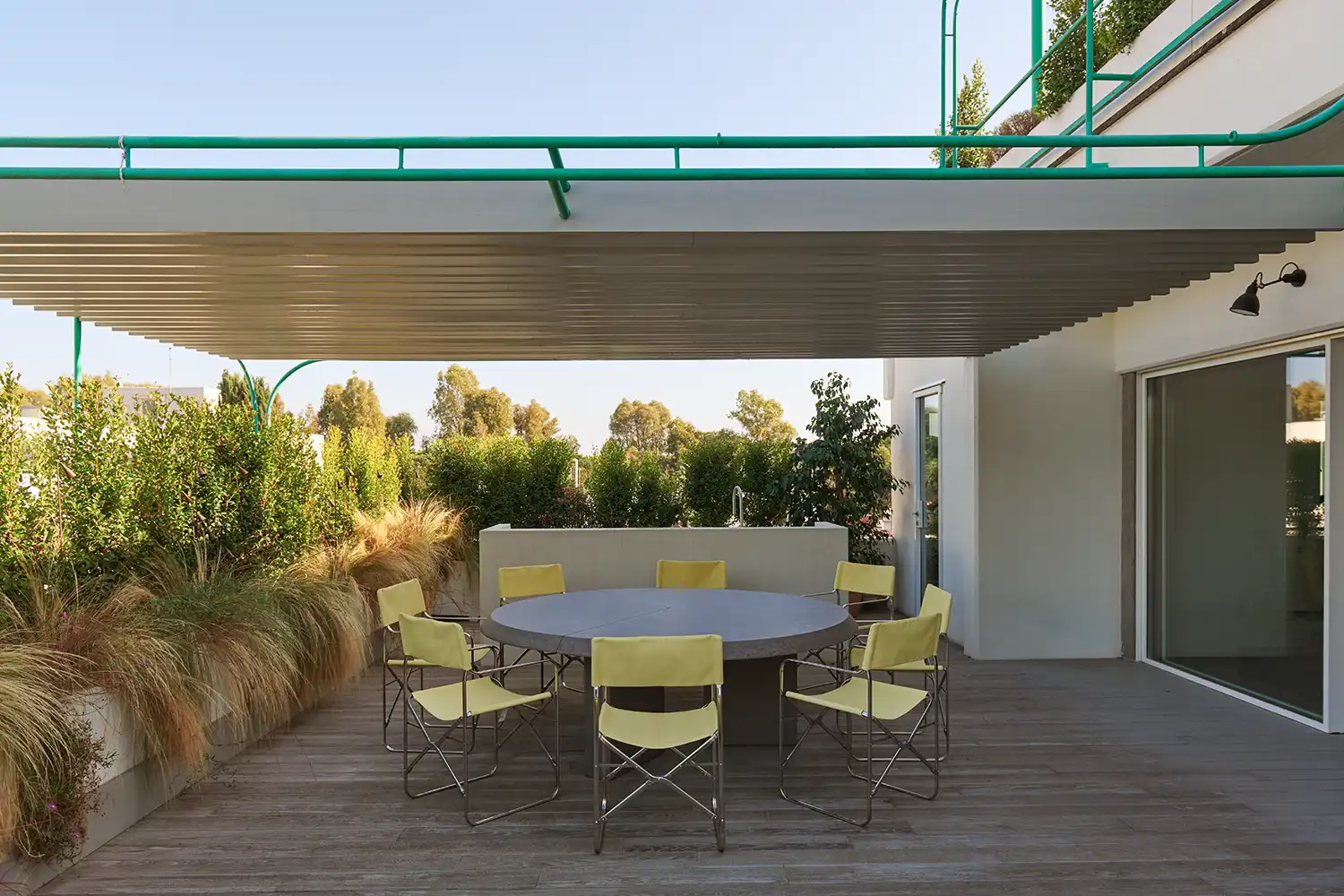 Outdoor terrace with a circular stone table, yellow Gae Aulenti chairs, and an aqua-green pergola.