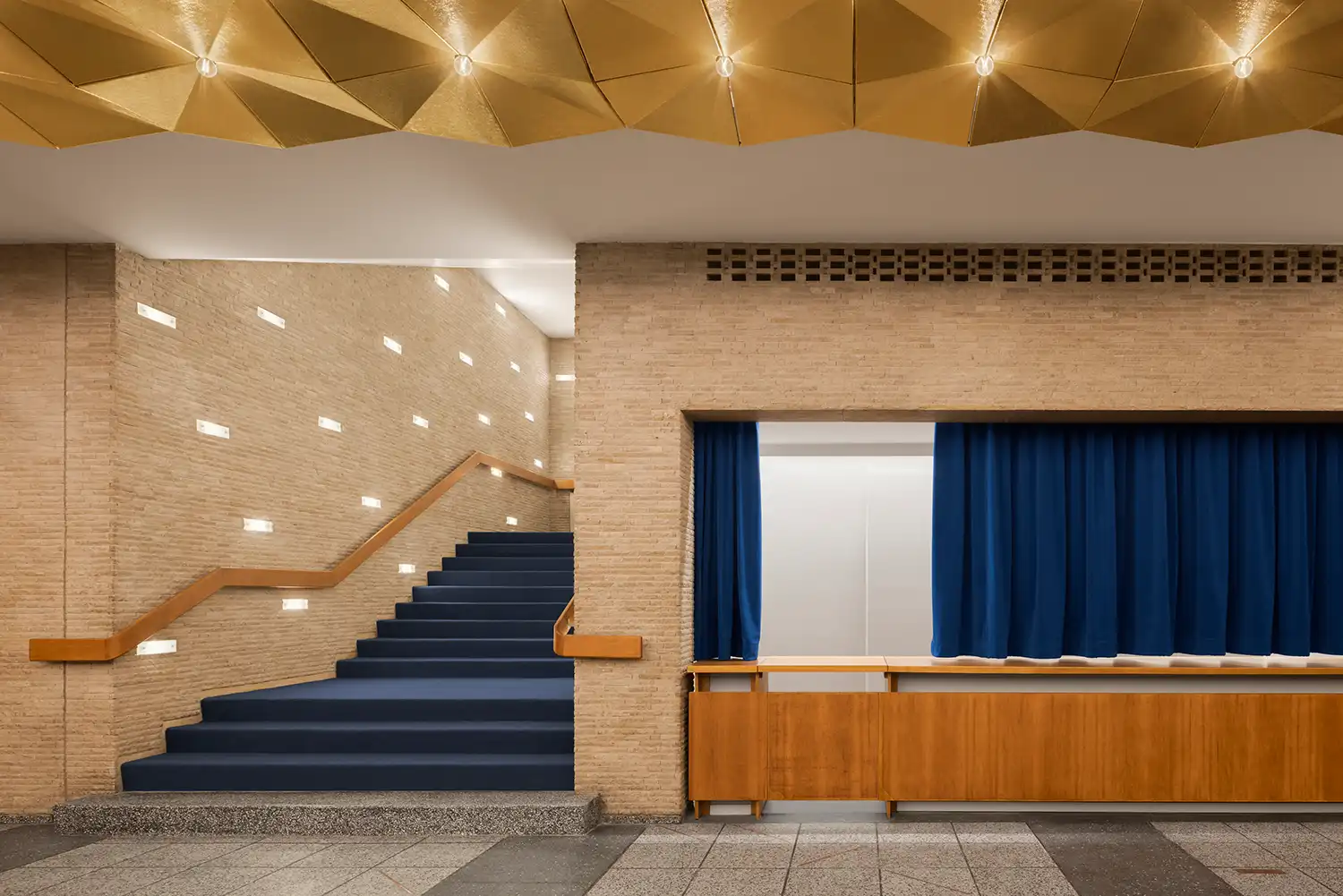Internal staircase with blue carpeting and wooden handrails beneath a geometric gold ceiling.