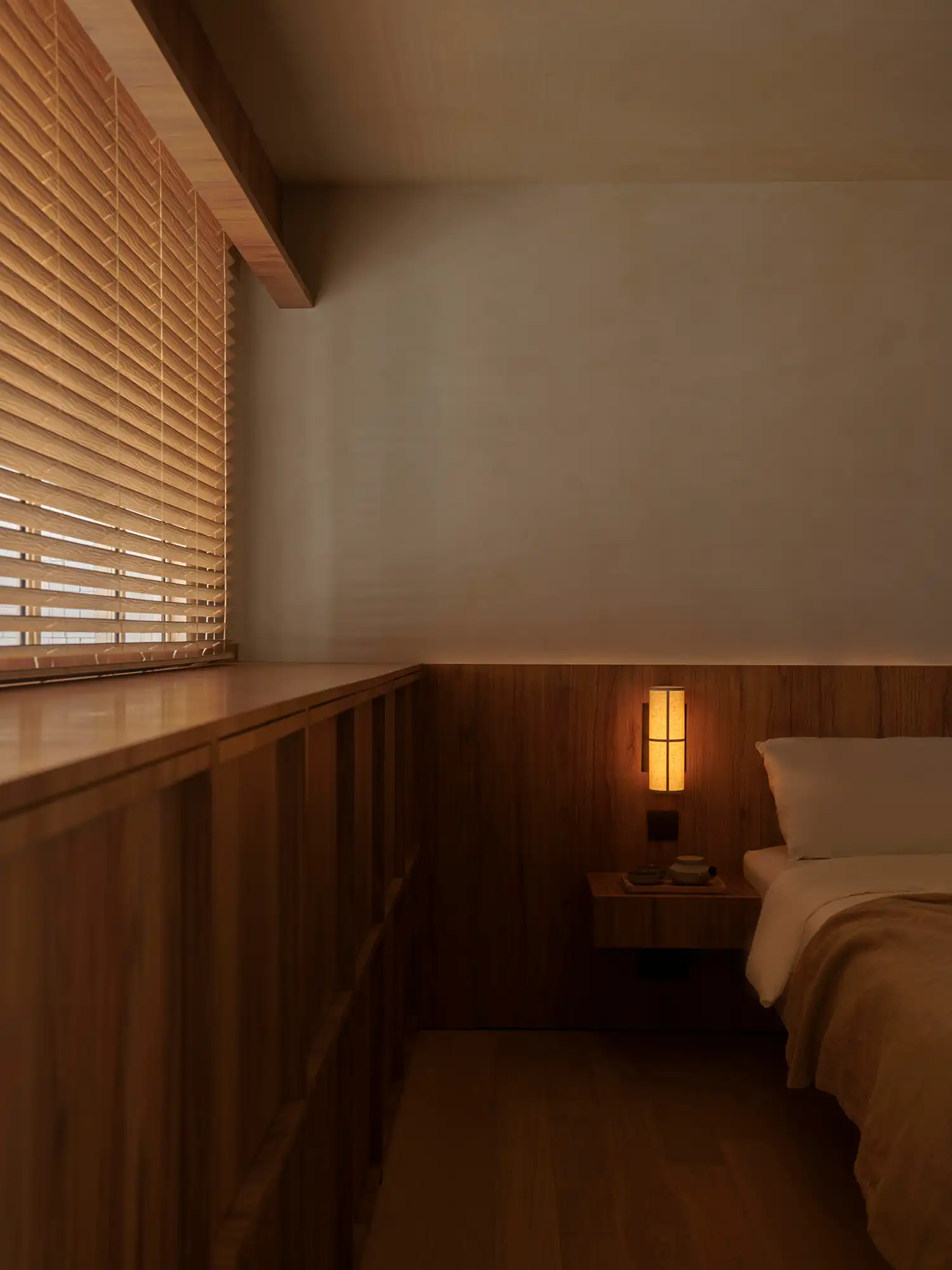 Minimalist bedroom with wood paneling, a warm wall lamp, and bamboo window blinds.
