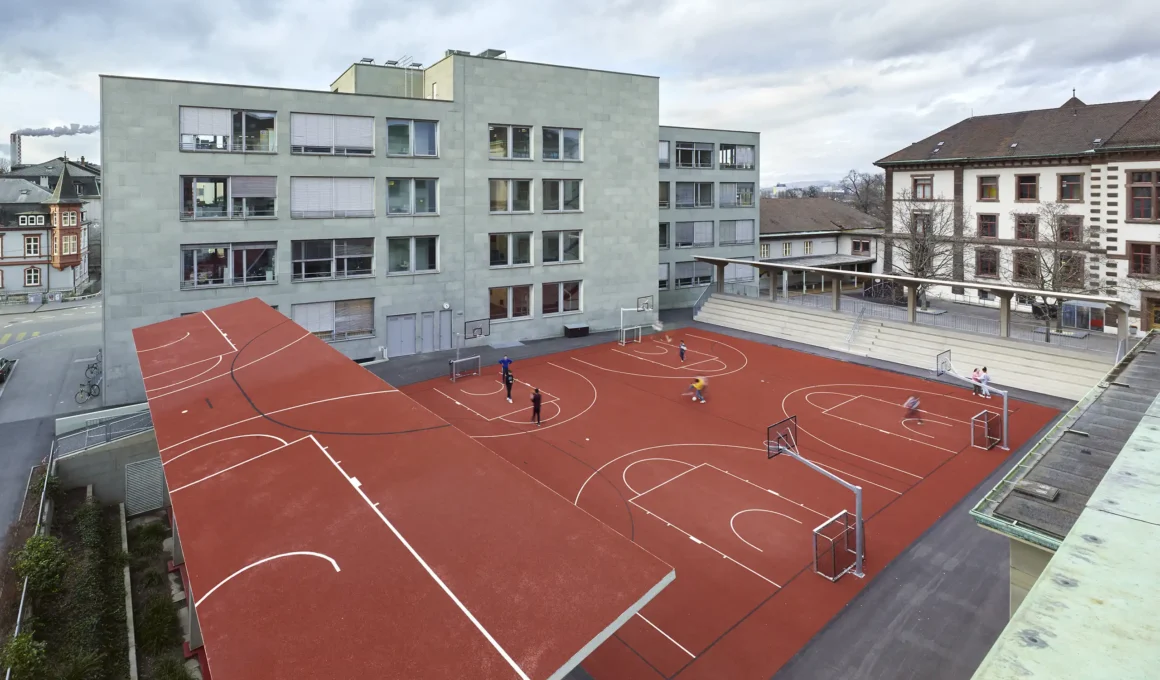 Wide aerial view of the red Vogesen stadium in Basel with students playing.