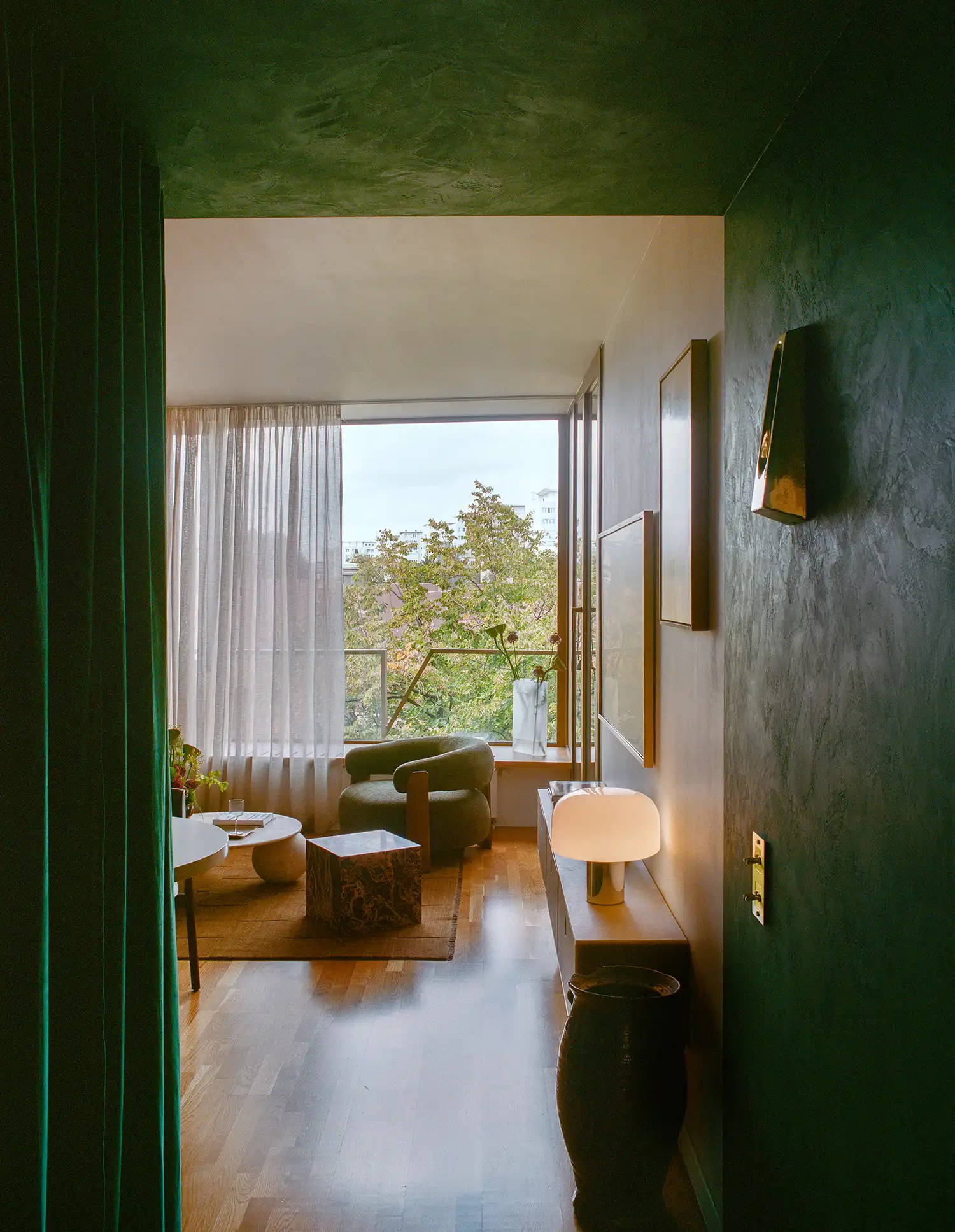 Perspective view from a dark green hallway looking into a sunlit living room with a historic tree outside.