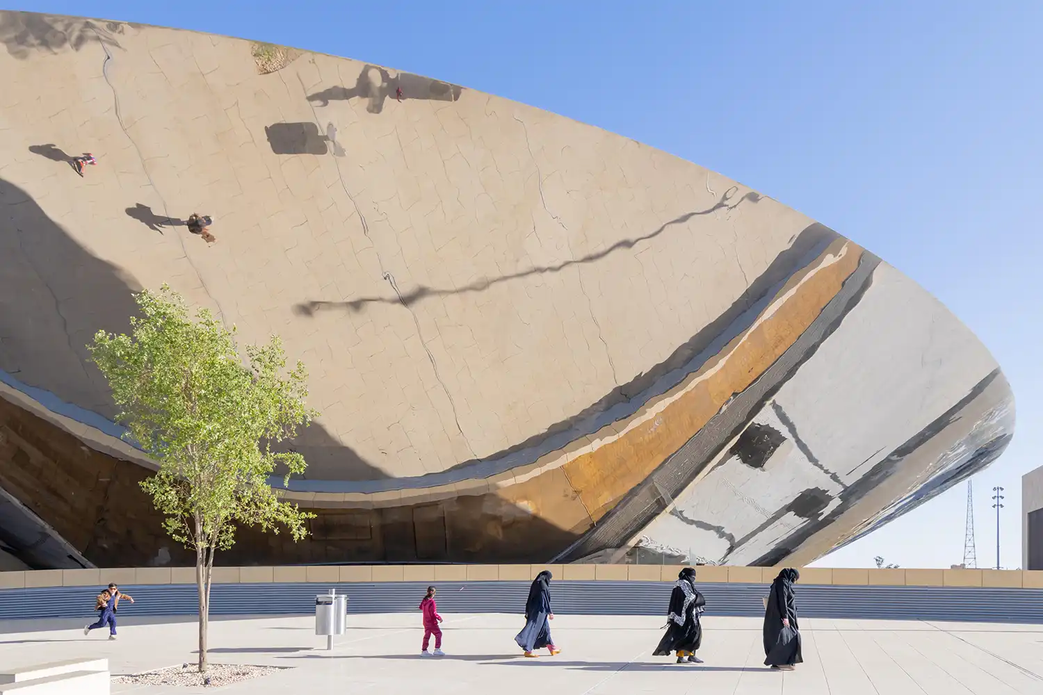 A low-angle shot of the massive polished steel canopy reflecting people walking on the light-colored plaza below.