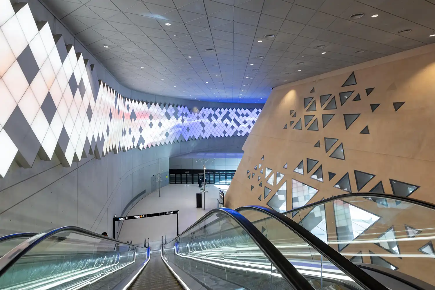 An interior view from an escalator looking down into the station towards a large media art wall with diamond patterns.