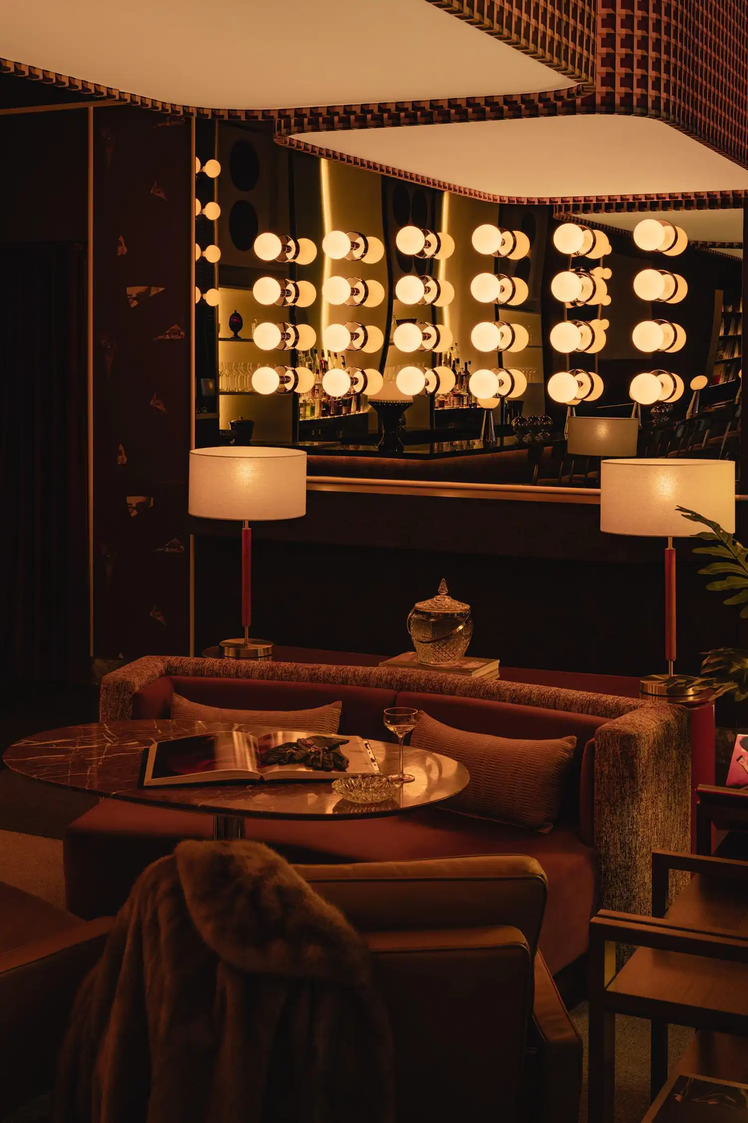 Sunken seating nook in Mimi with a marble table, velvet sofa, and warm table lamps against a mirrored wall.