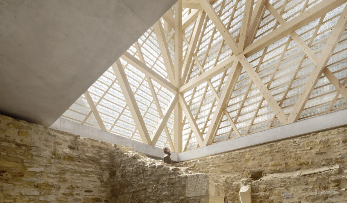 Interior view of medieval stone walls meeting a modern timber truss ceiling with glass tiles.