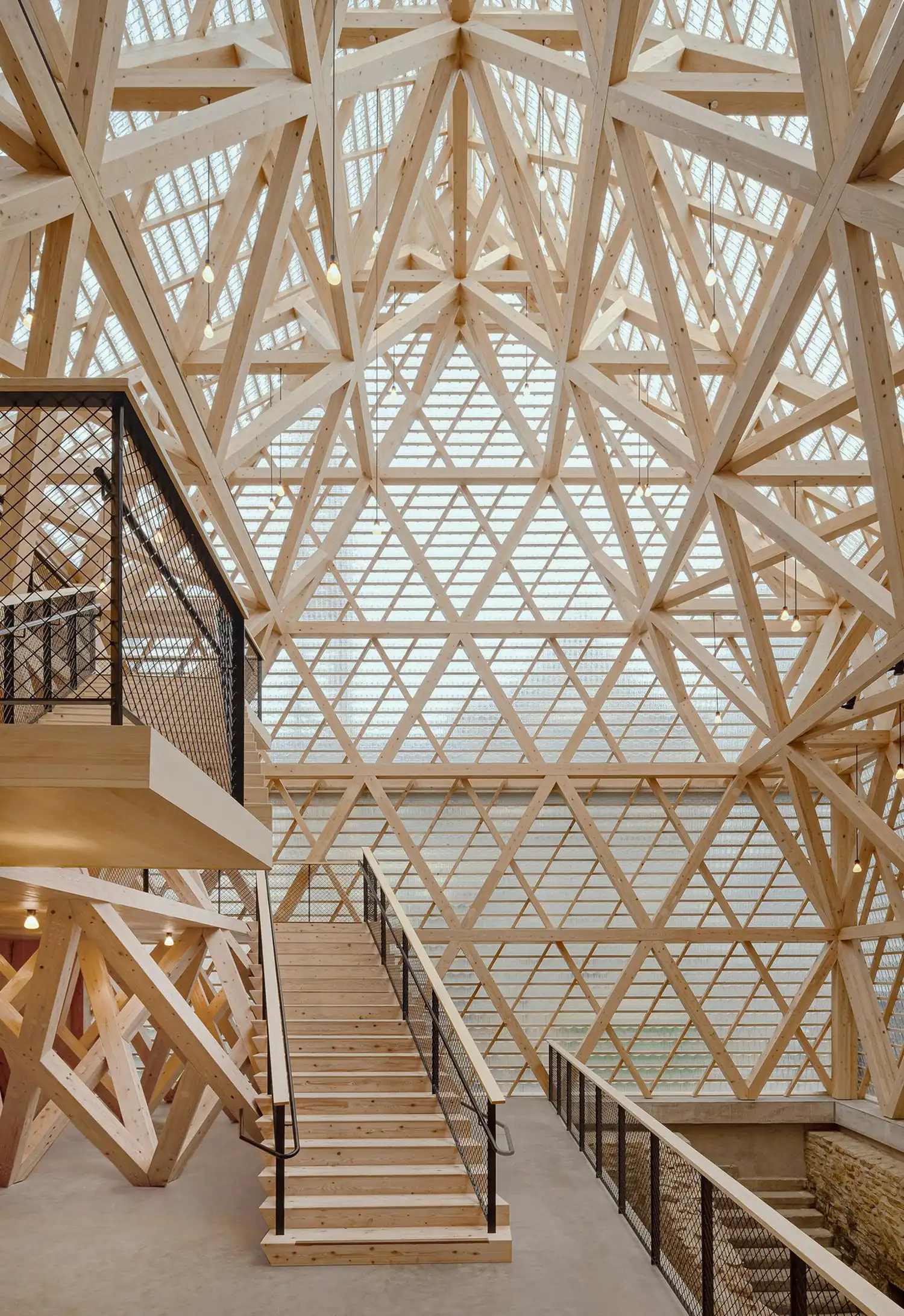 Interior view of a multi-story atrium with a complex wooden truss system and a wooden staircase.