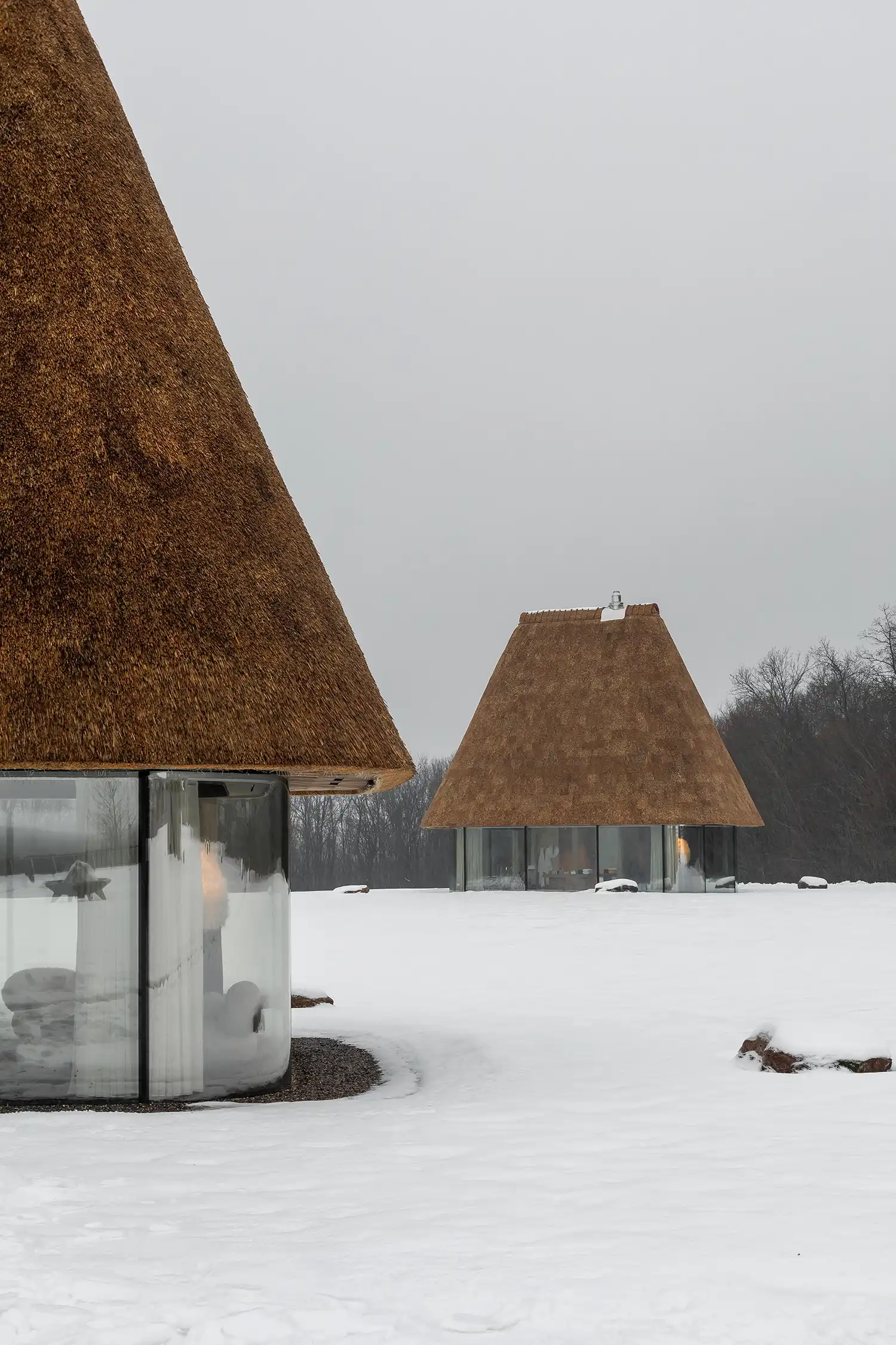 Two glass guesthouses with oversized thatched roofs in a snowy landscape.
