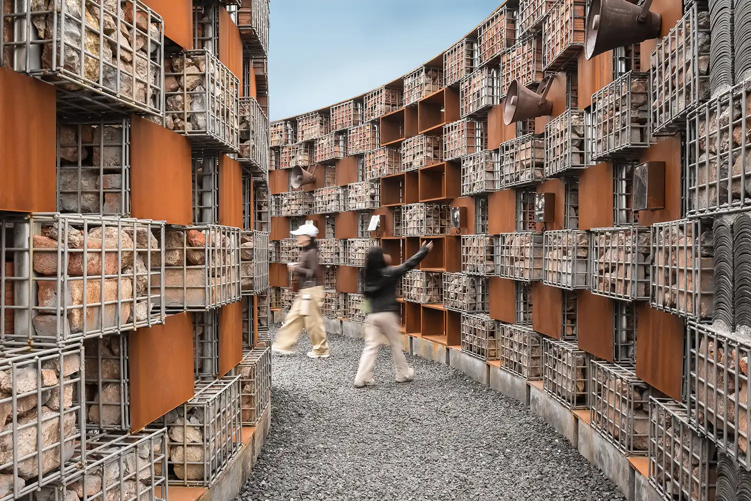 Visitors walking through the narrow gravel path between gabion walls and metal megaphones.