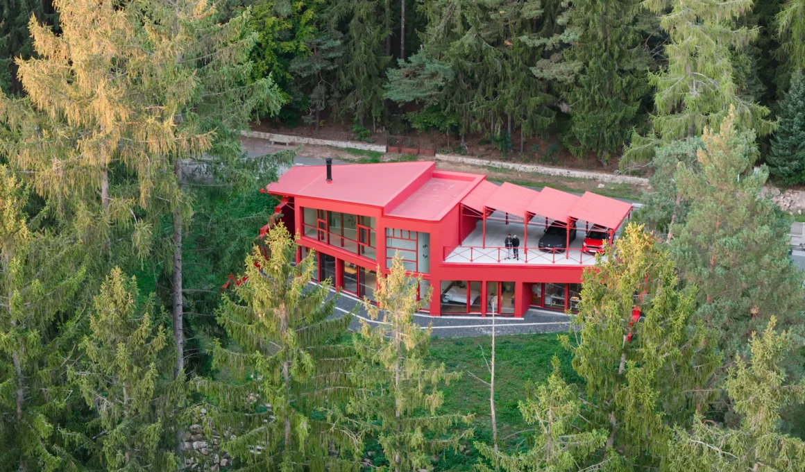 Aerial view of a bright red two-story villa with a sawtooth roof nestled in a dense green forest.