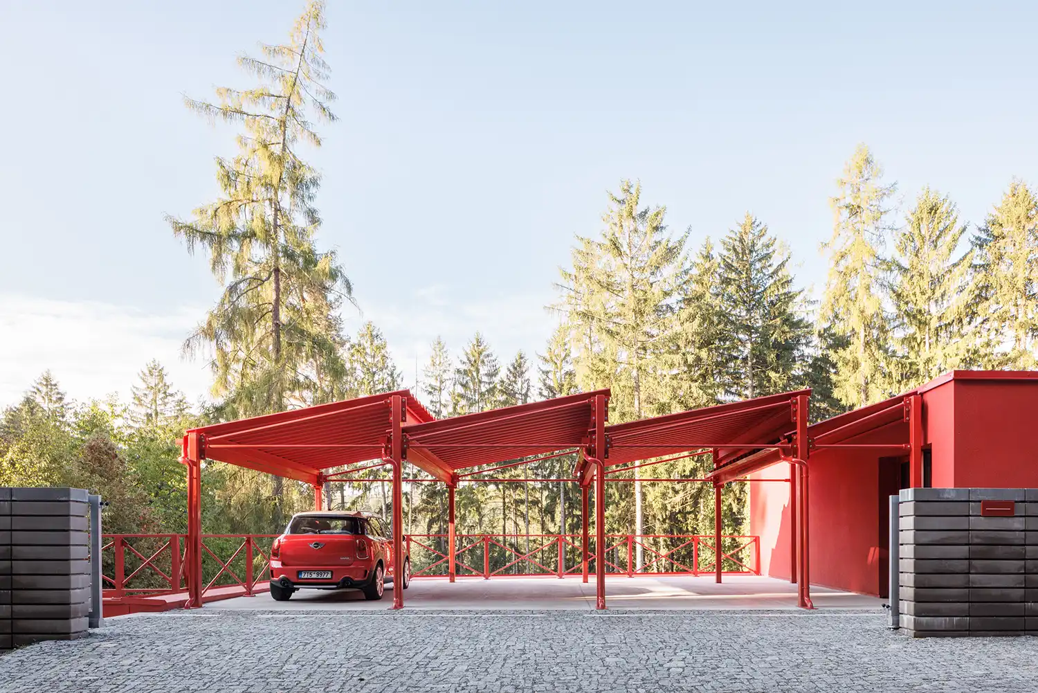 Red steel sawtooth carport structure with a red car parked on a stone-paved entrance.
